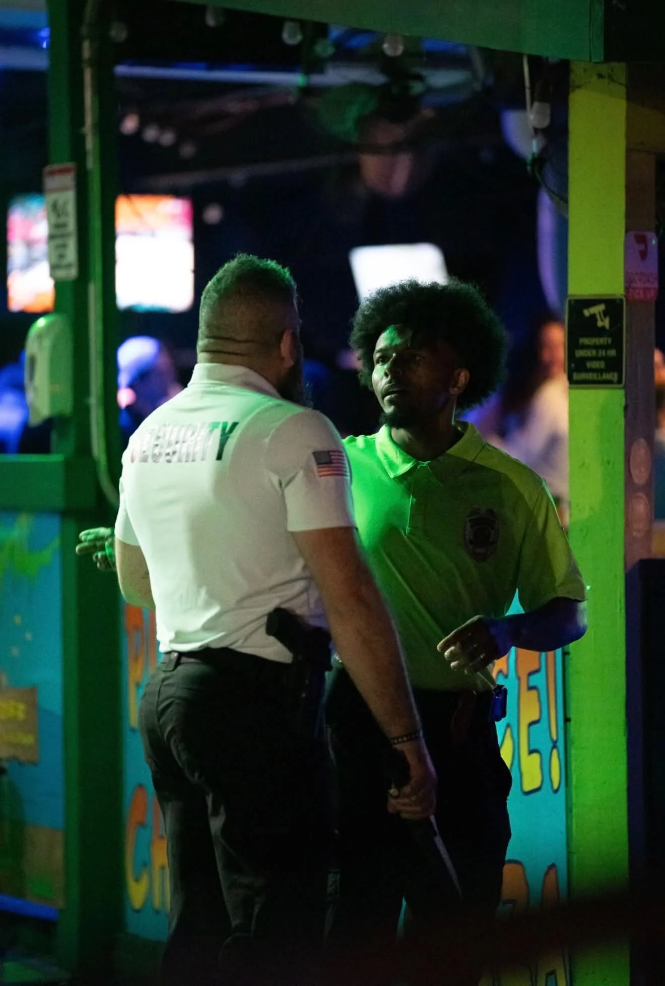 Two men, one in a security uniform and one in a green shirt, talking at night near a bright, colorful amusement park or arcade entrance illuminated with neon lights.