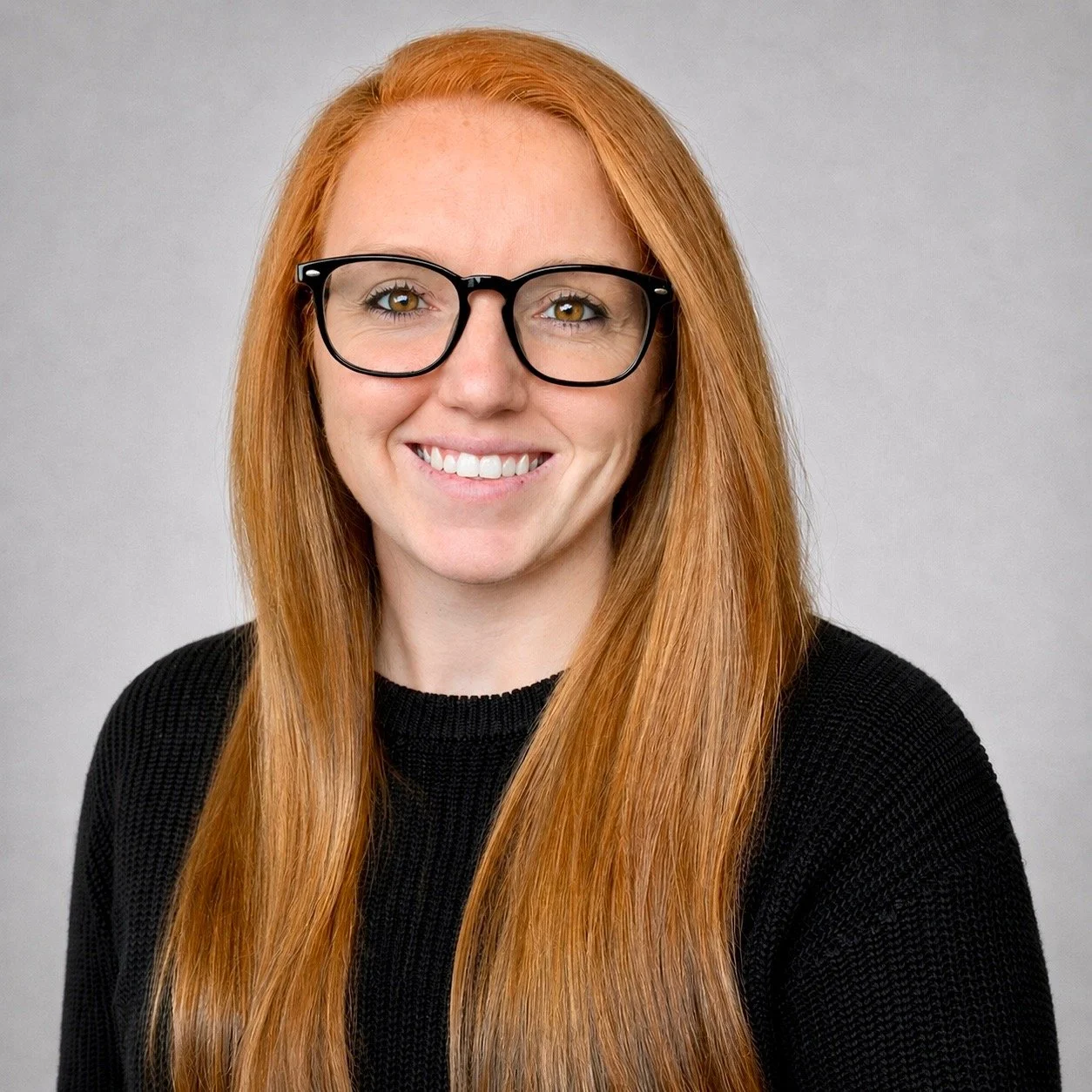 Portrait of a smiling woman with long red hair, wearing glasses and a black shirt, against a plain gray background.
