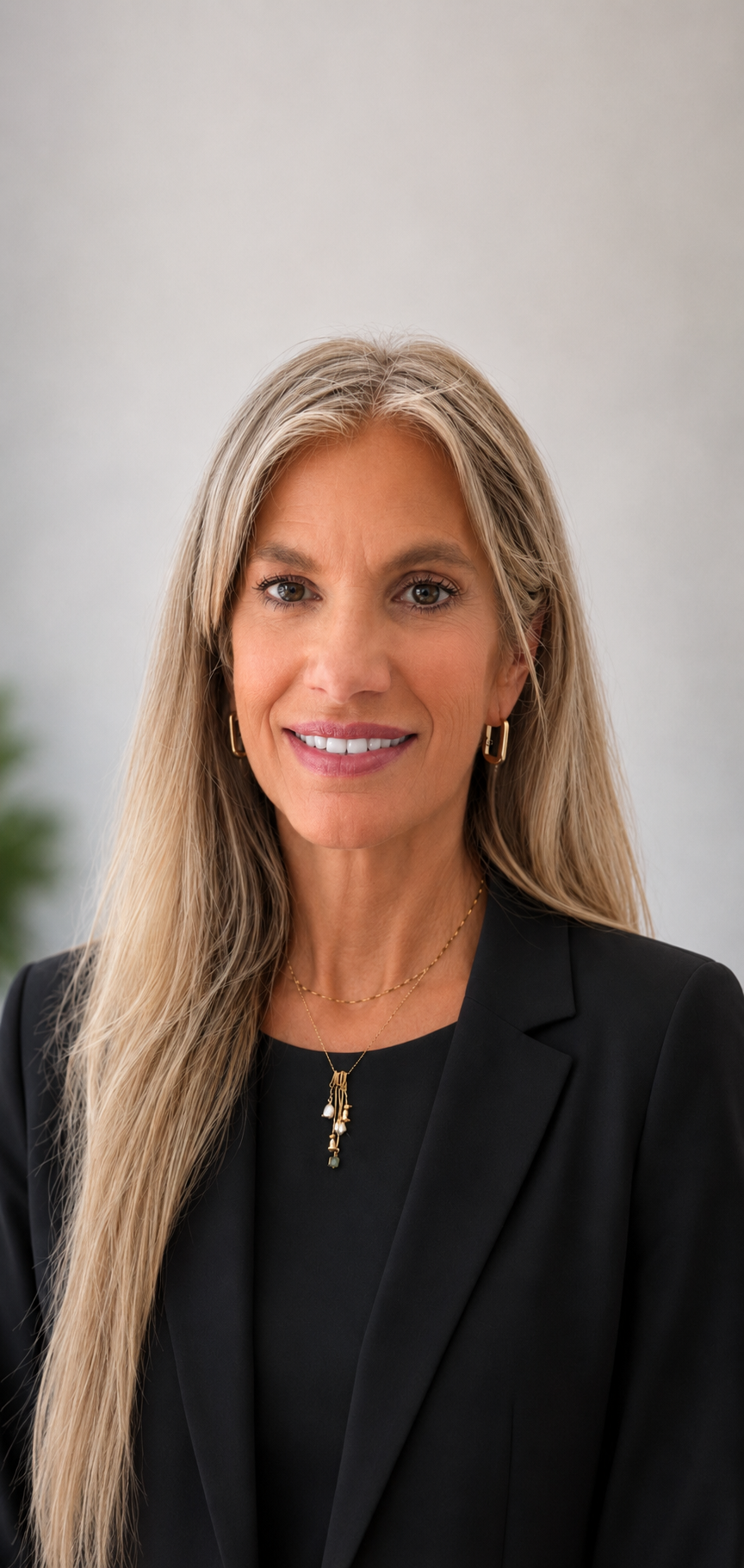 A smiling woman with long blonde hair wearing a black blazer, gold jewelry including earrings, a necklace, and a pendant, against a neutral background.