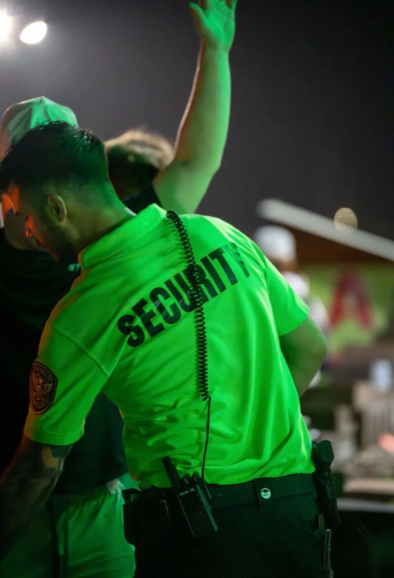 A police officer wearing a bright yellow safety vest with the word 'SECURITY' on the back is standing at night. The officer has a radio attached to their belt and appears to be engaged in some activity or conversation. The background is dark with indistinct lights.