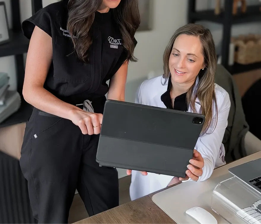 Two women in an office, one sitting and one standing, looking at a tablet. The sitting woman is smiling, and the standing woman is pointing at the tablet. There is a laptop and a mouse on the desk.