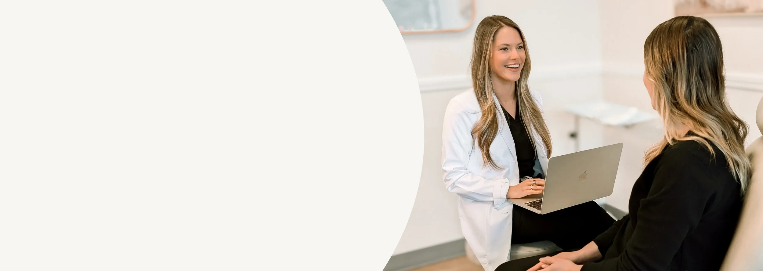A woman in a white coat with a laptop talking to another woman sitting in a chair in a medical office.