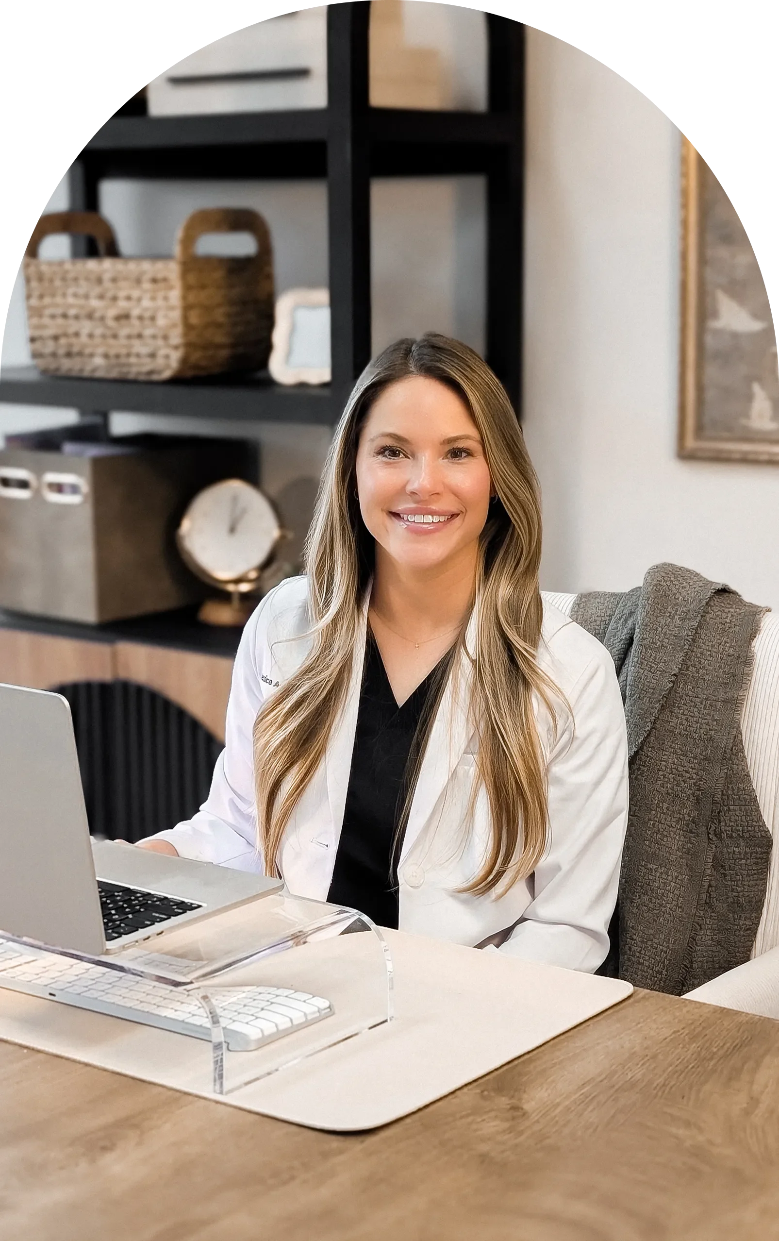 A smiling woman with long, wavy blonde hair, wearing a white coat, sitting at a desk with a laptop and a wireless keyboard, in a modern office or workspace with shelves, a clock, and artwork in the background.