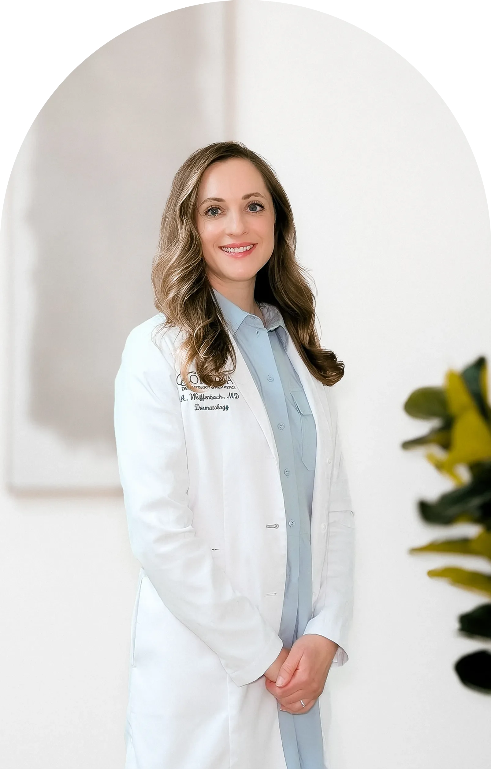 A woman with long wavy hair wearing a white lab coat over a light blue shirt, smiling, standing against a plain white background.