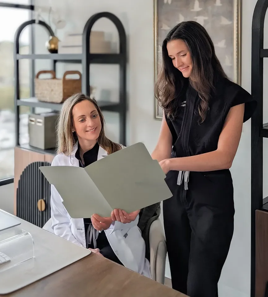 Two women, one in a white lab coat and the other in a black uniform, looking at a clipboard together in an office setting.