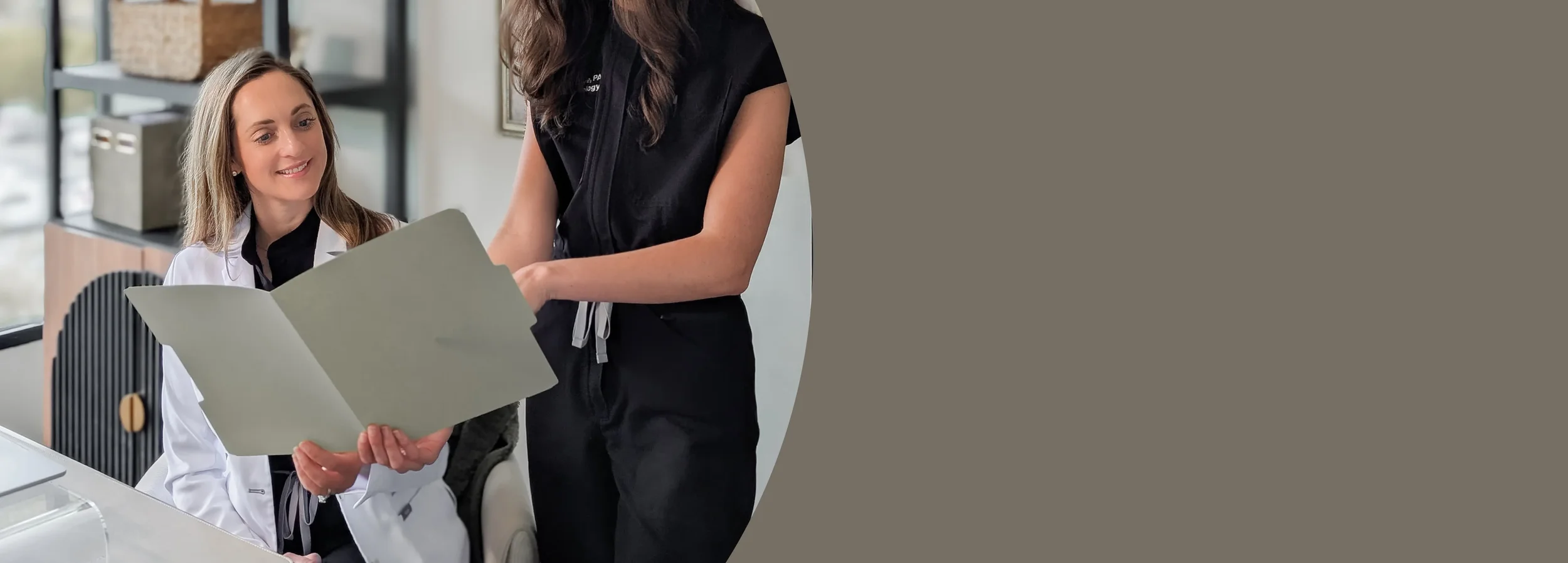 Two women in an office, one seated with a white coat, the other standing in a black outfit, looking at a folder together.