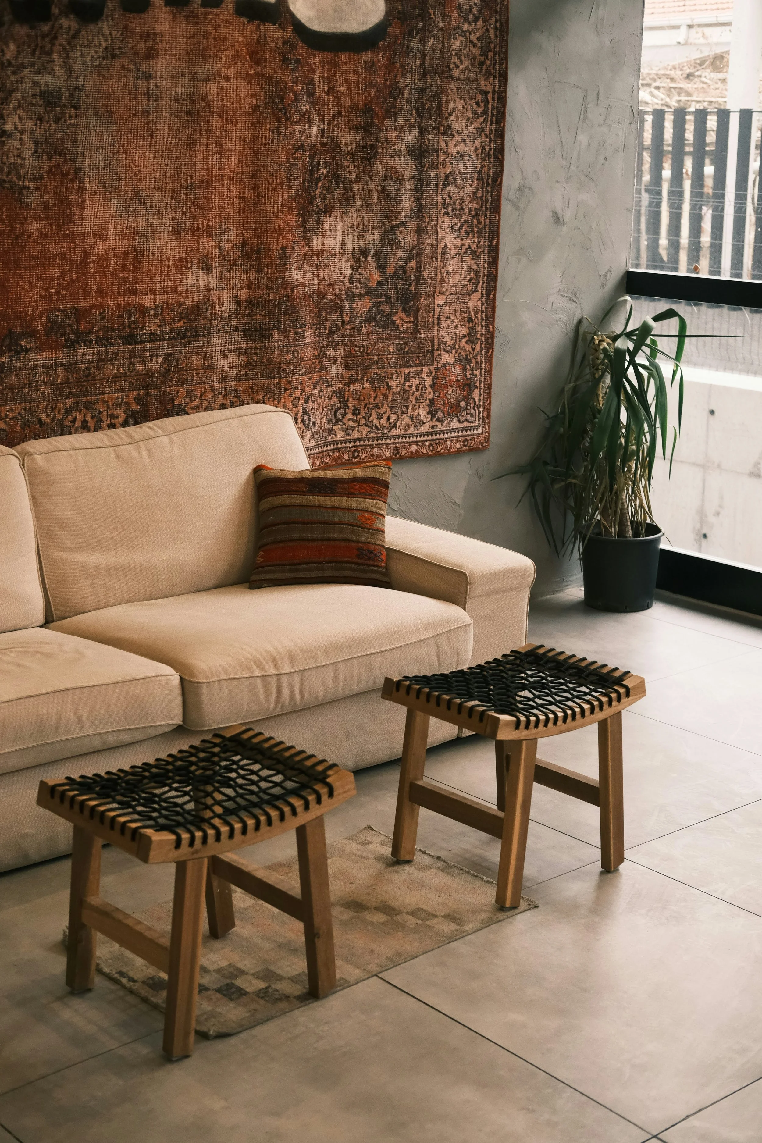 A cozy indoor living space featuring a cream-colored sofa with a striped pillow, a vintage rug, a large potted plant, wooden stools with woven seats, and a window with a black frame letting in natural light.