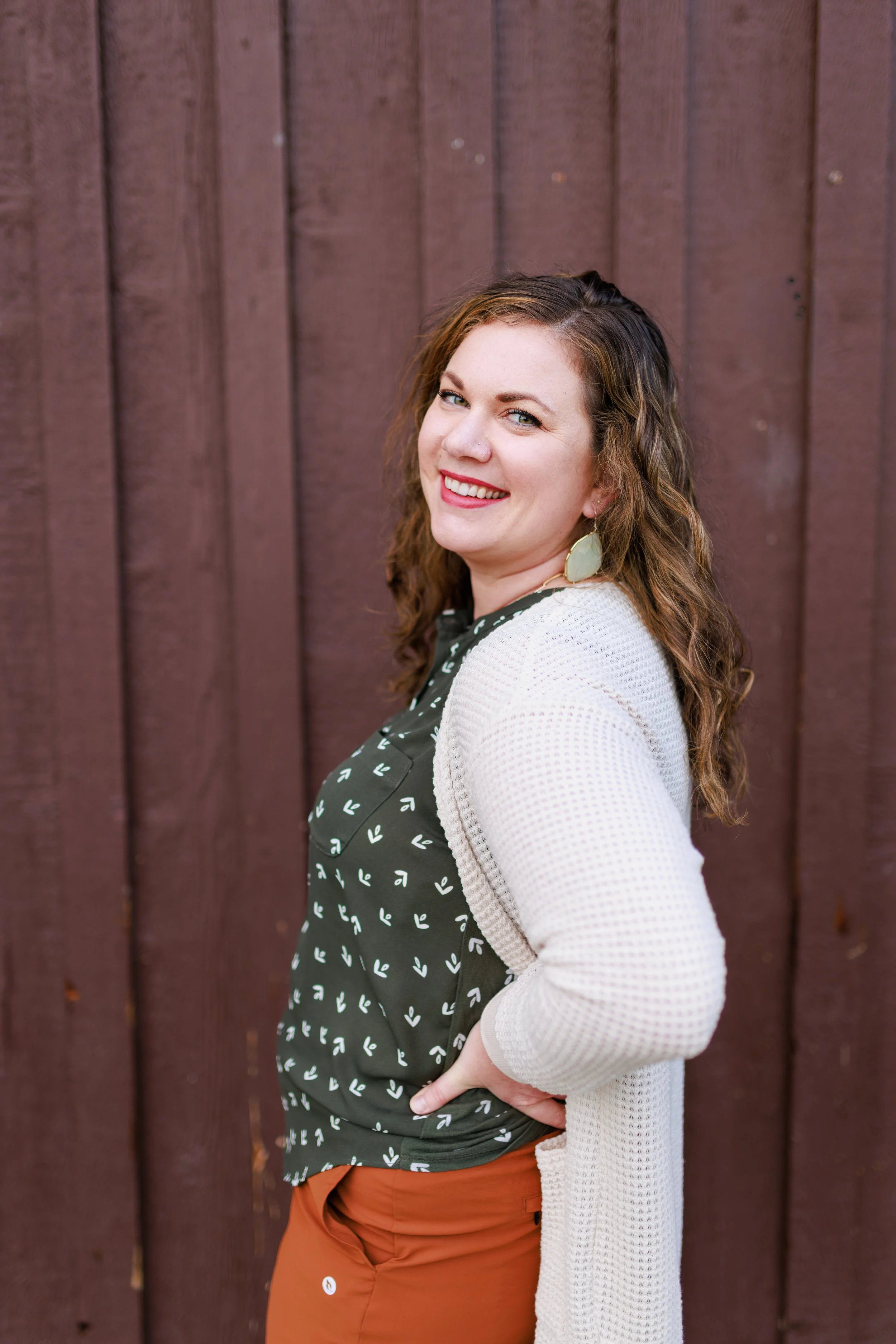 Smile and upper body of a woman with wavy brown hair, wearing green earrings, a black patterned top, a white cardigan, and rust-colored pants, standing against a wooden fence.