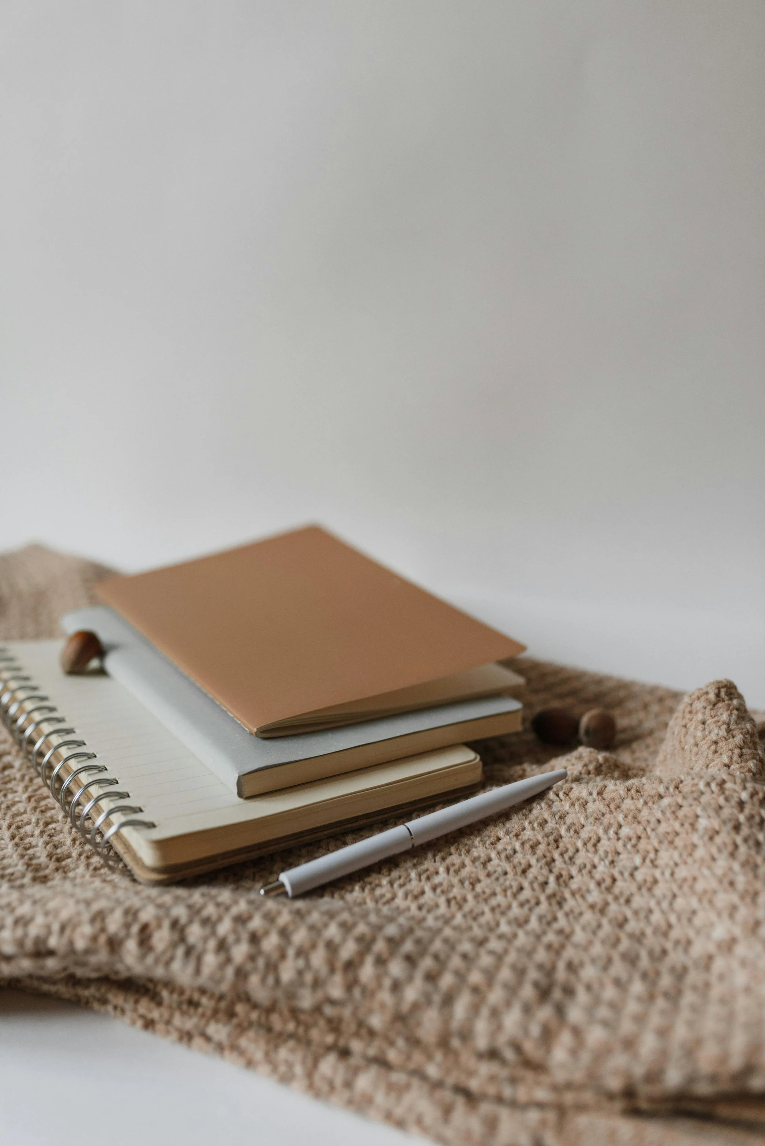 Stacked notebooks, a white pen, and acorns placed on a textured beige carpet.