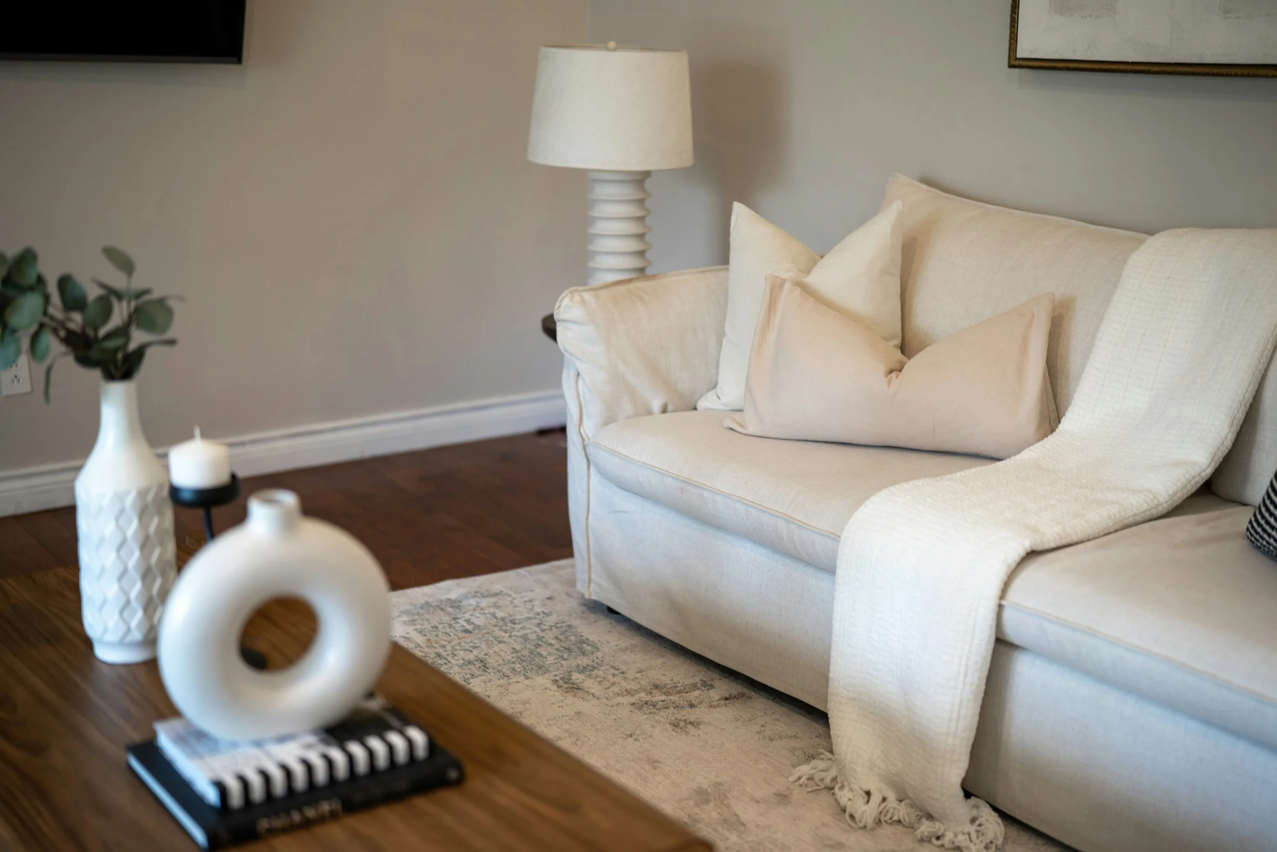 Living room with a beige sofa, white pillows, a white table lamp, and decorative vases on a wooden table.