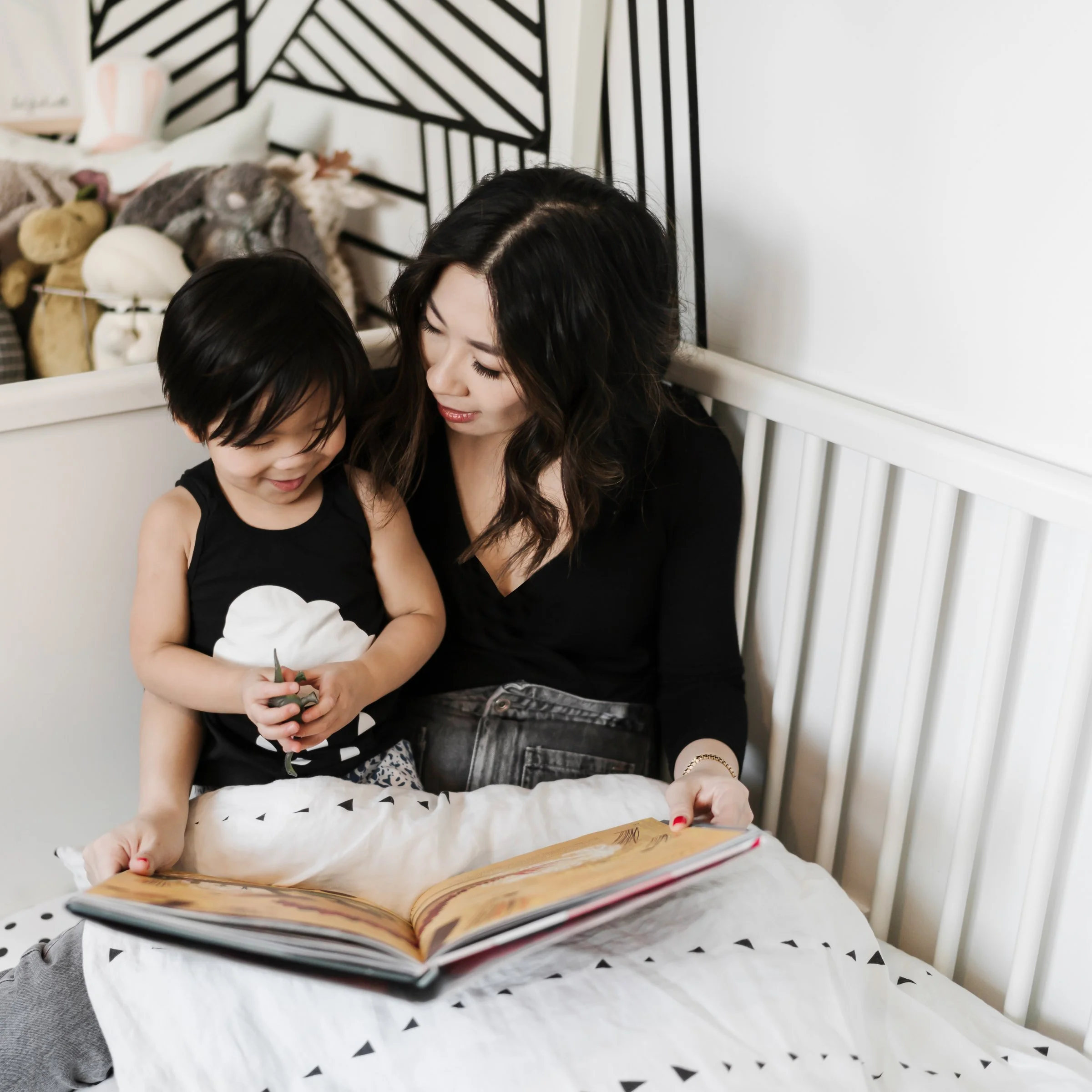 A woman and a young girl sitting on a bed, looking at a book together, with a pile of stuffed animals and pillows in the background.