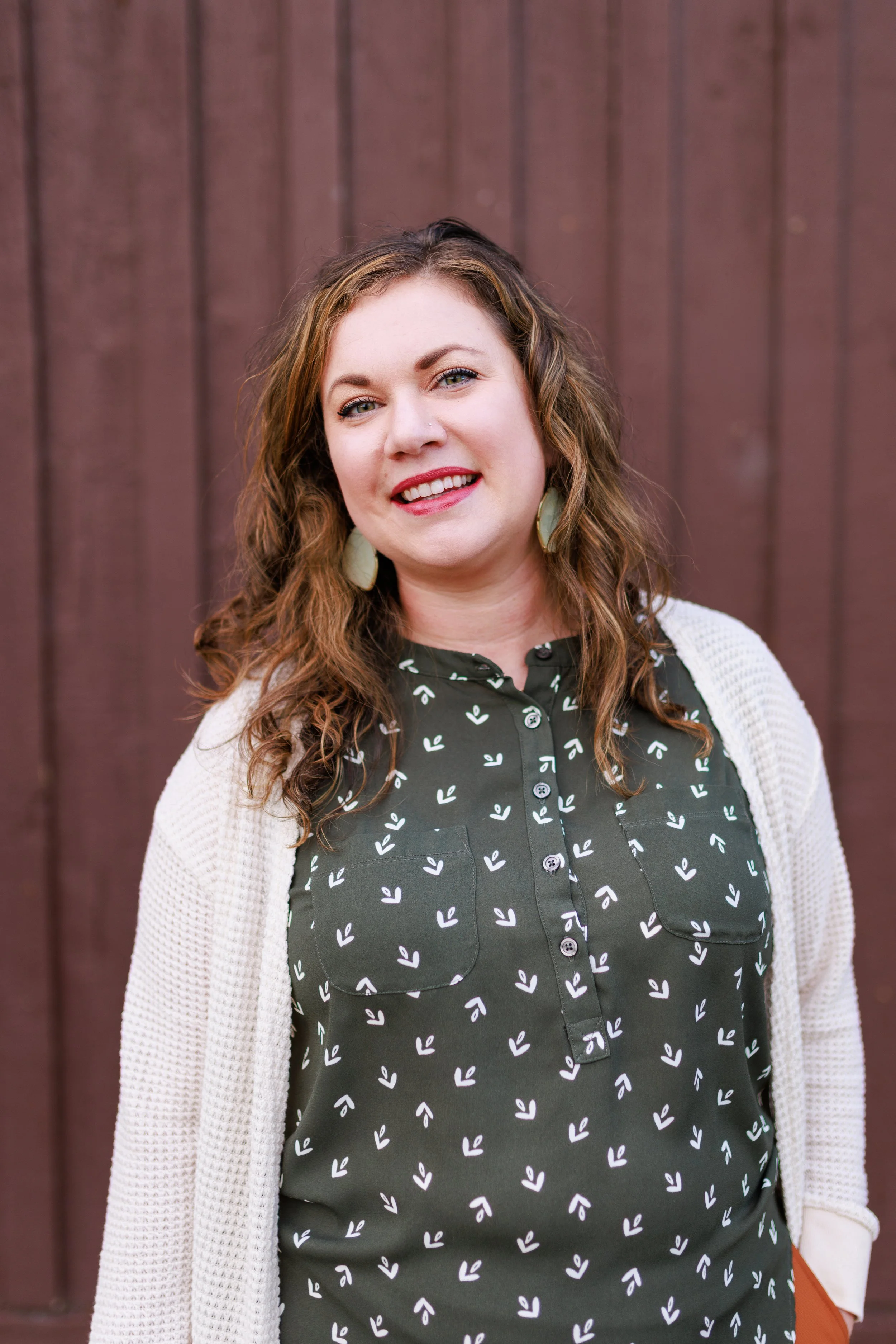 A woman with wavy brown hair and light skin is smiling in front of a brown wooden wall. She is wearing a black shirt with a white leaf pattern and a beige cardigan.