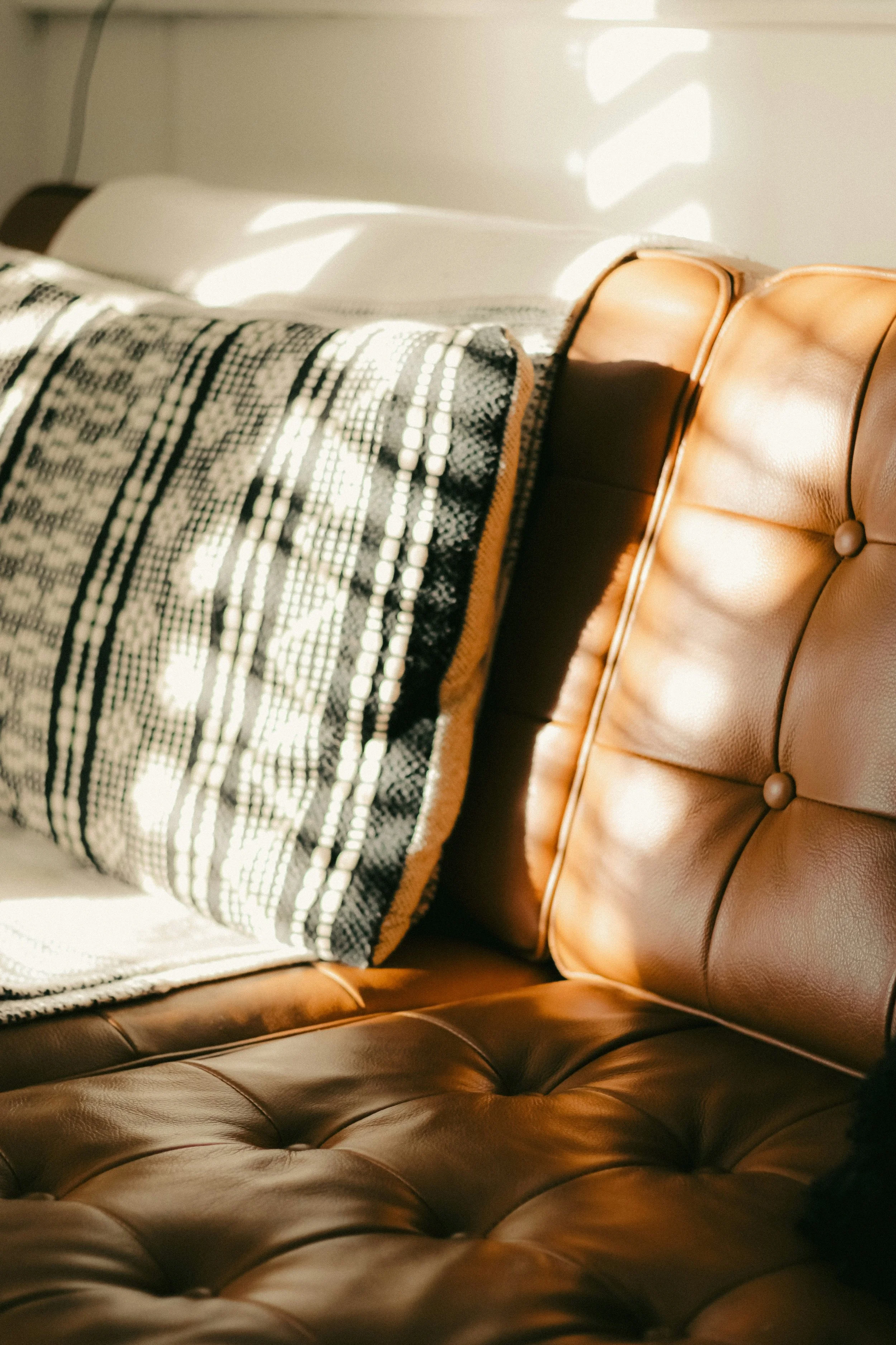 Close-up of a brown leather tufted sofa with sunlight casting shadows on it, and a patterned blanket draped over the backrest.