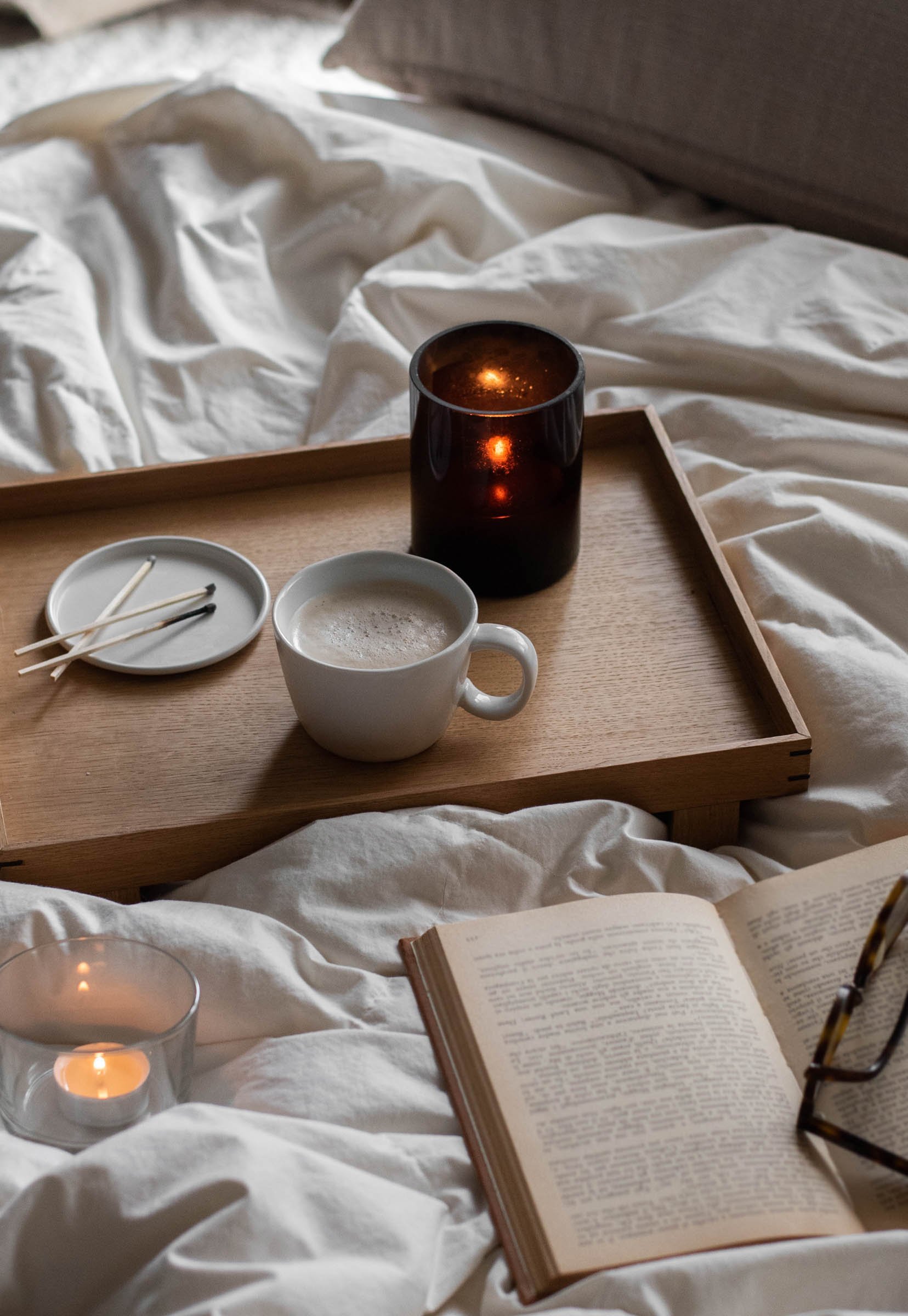 A wooden breakfast tray with a white mug of coffee, a small dish with toothpicks, a lit candle, and a candle holder, placed on a white bed with an open book and a pair of glasses nearby.