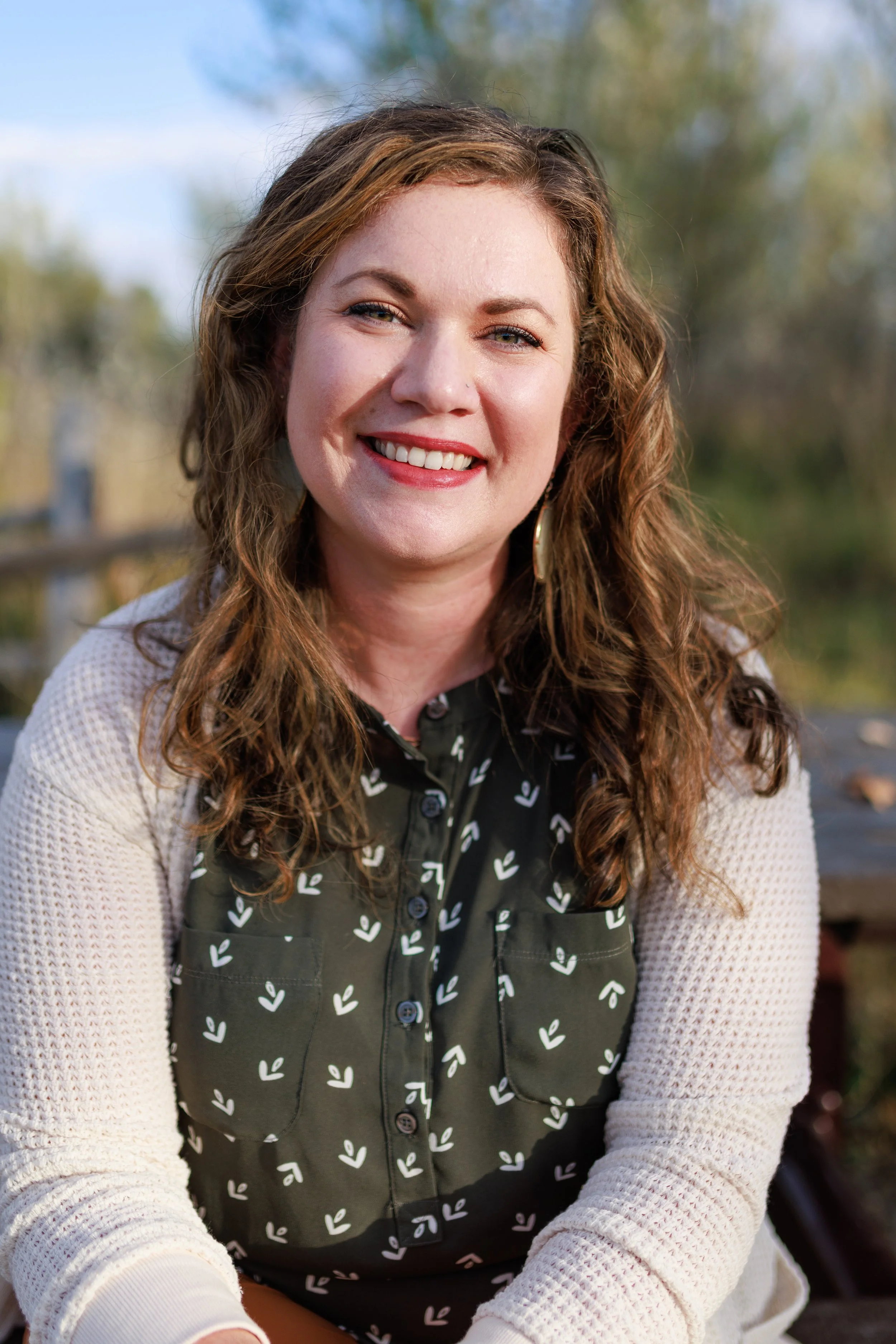 Smiling woman with curly red hair, wearing a black shirt with white leaf patterns and a beige cardigan, sitting outdoors on a sunny day.