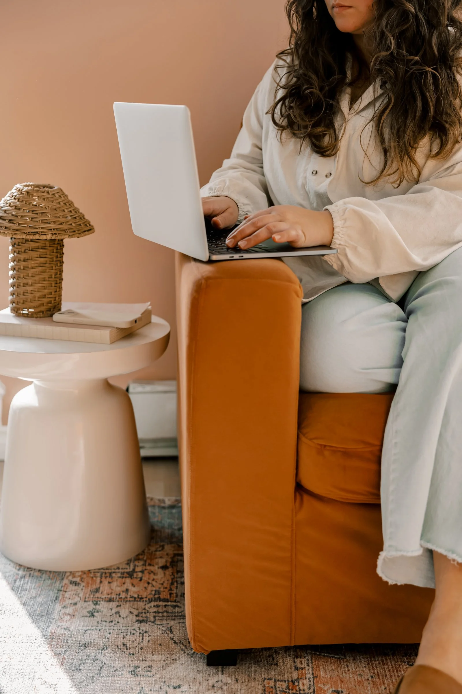 A woman with curly hair sitting on an orange sofa, working on a laptop. A small white side table with a woven lampshade, a closed notebook, and a smartphone is next to her. The room has warm tones and a patterned rug.