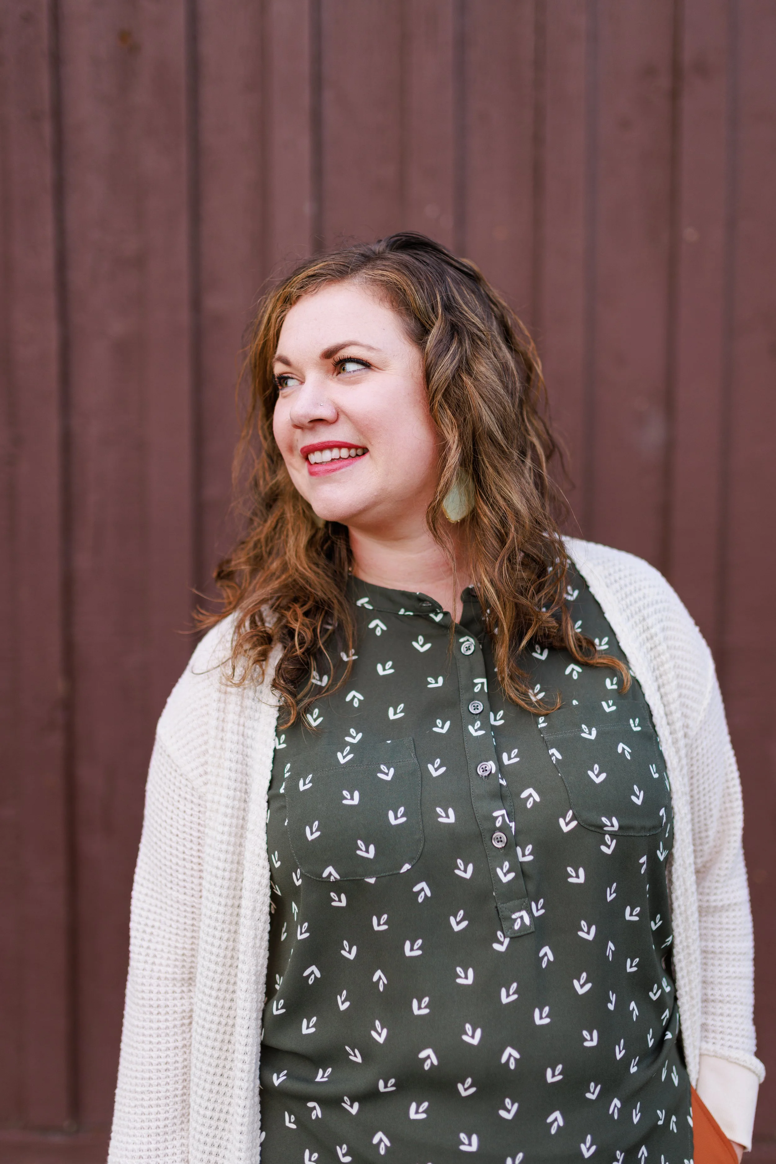 A woman with curly brown hair smiling, standing in front of a wooden wall, wearing a dark green dress with white leaf patterns and a white cardigan.