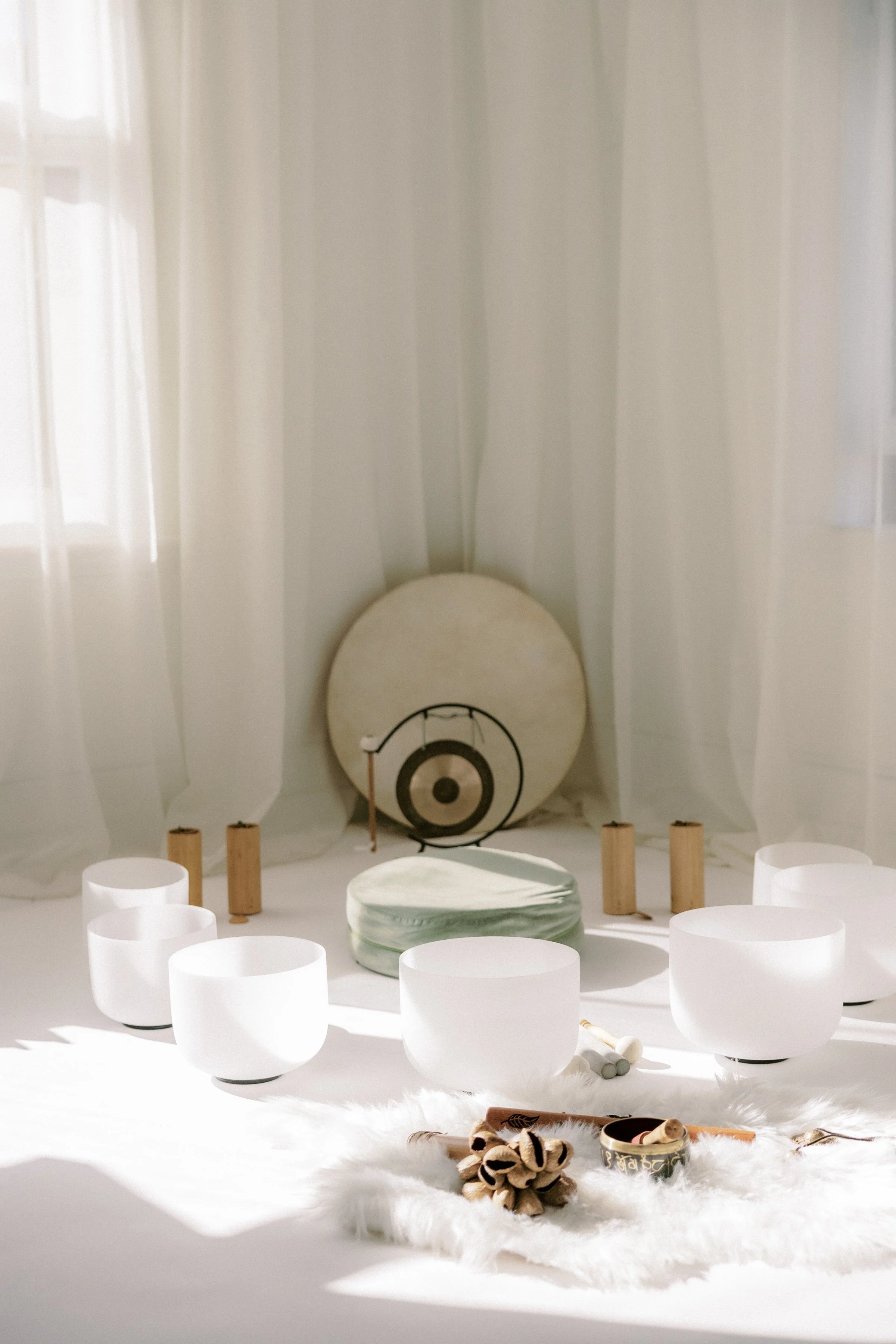 A white altar with a green cushion, surrounded by white candles, with a large drum in the background and small wooden objects and plush toys on a white furry cloth in the foreground. White curtains frame the scene.