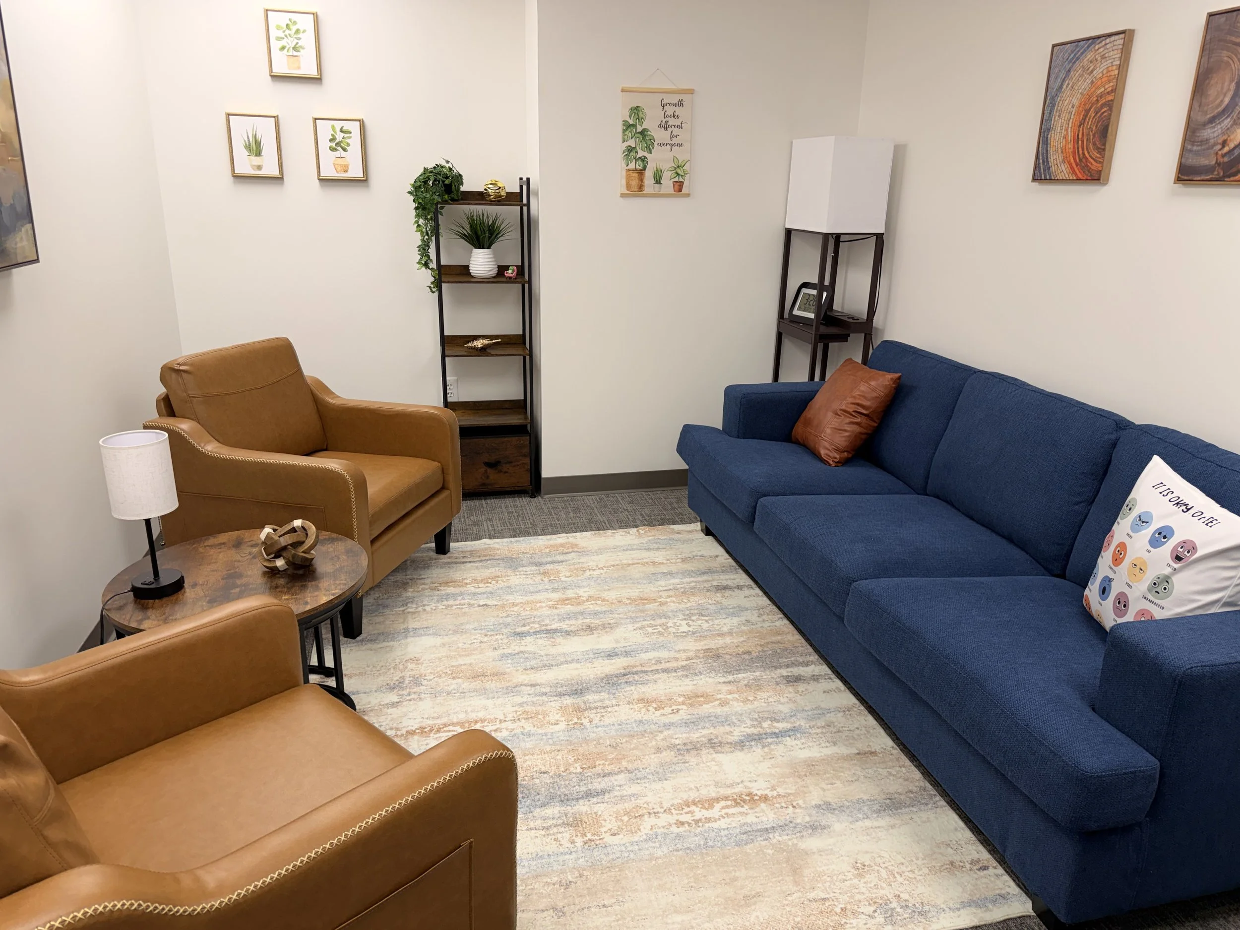 A small waiting room with beige, blue, and brown chairs, a wooden coffee table, a black metal shelf with plants and decorative objects, framed artwork of plants on the wall, a whiteboard, and colorful abstract paintings, with a colorful striped rug on the floor.