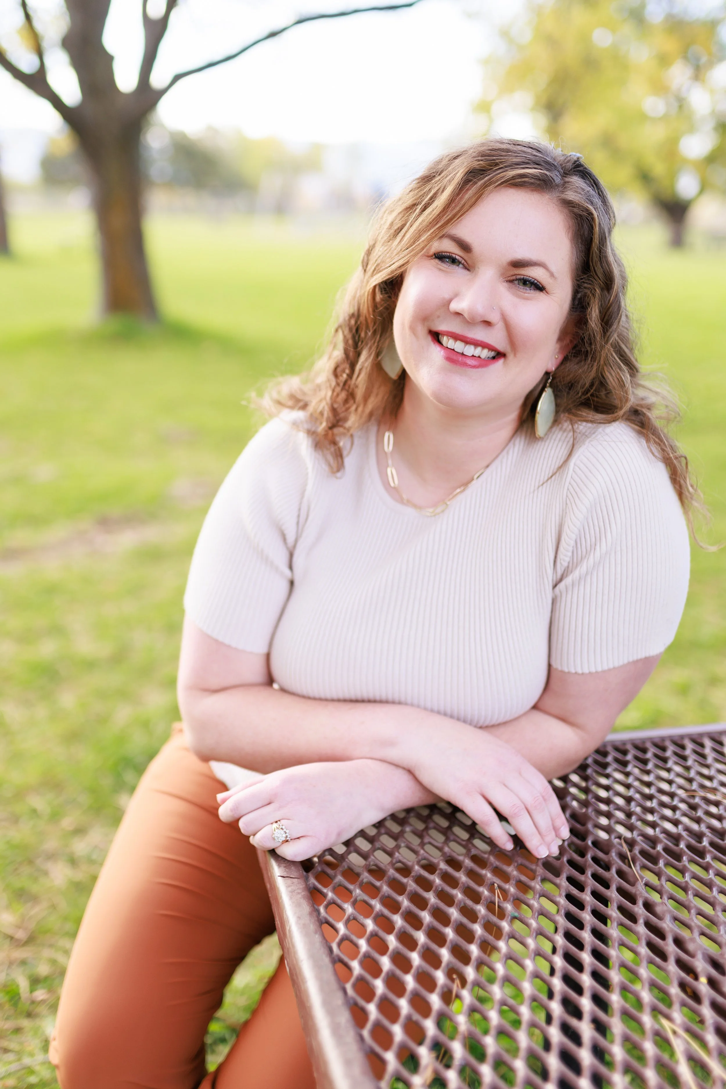 A woman with curly light brown hair, wearing a cream-colored short-sleeve top and brown pants, sitting outdoors at a picnic table, smiling with arms crossed, in a park with green grass and trees.