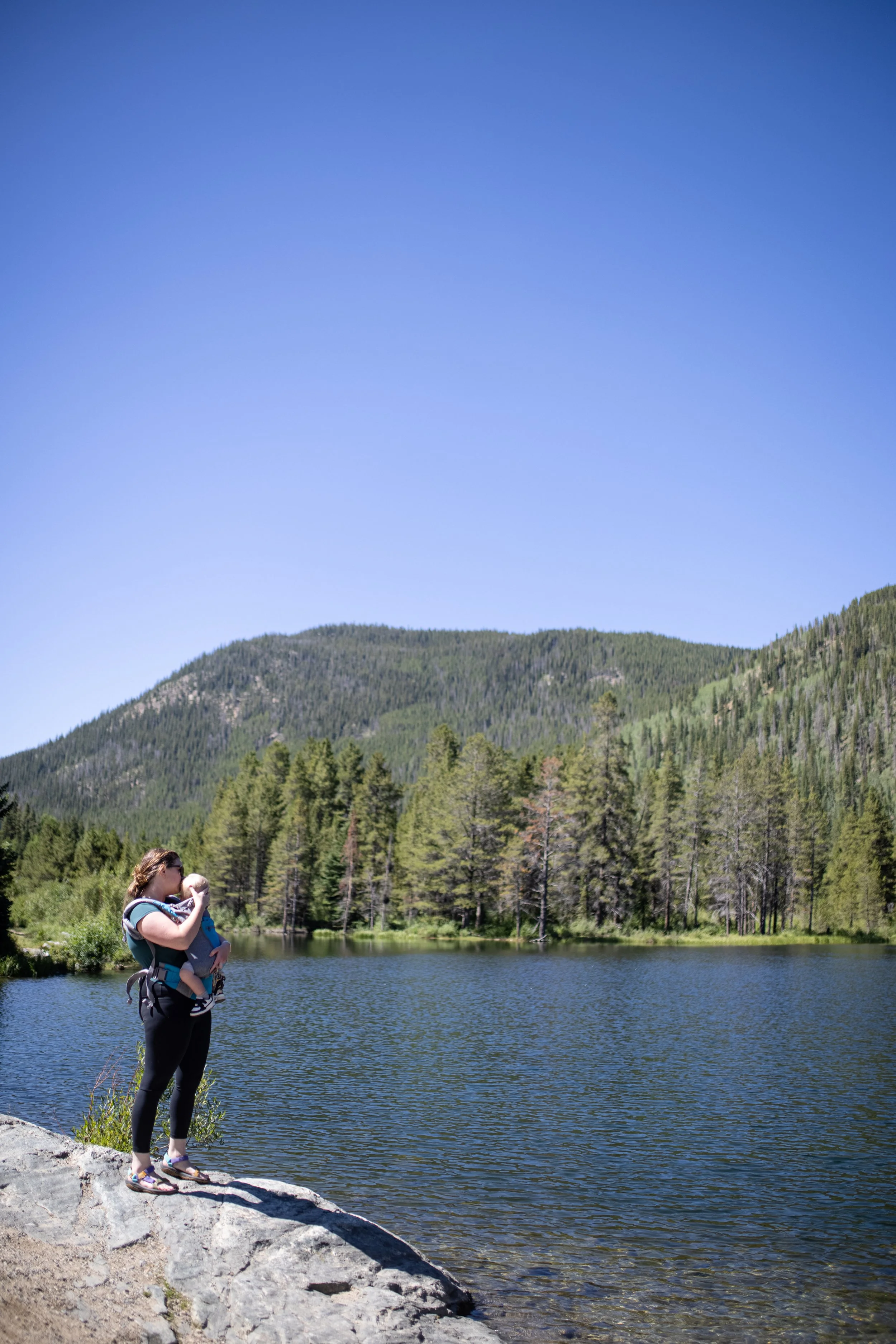 A woman carrying a child on her chest stands on a large rock by a lake, surrounded by trees and mountains, with a clear blue sky overhead.