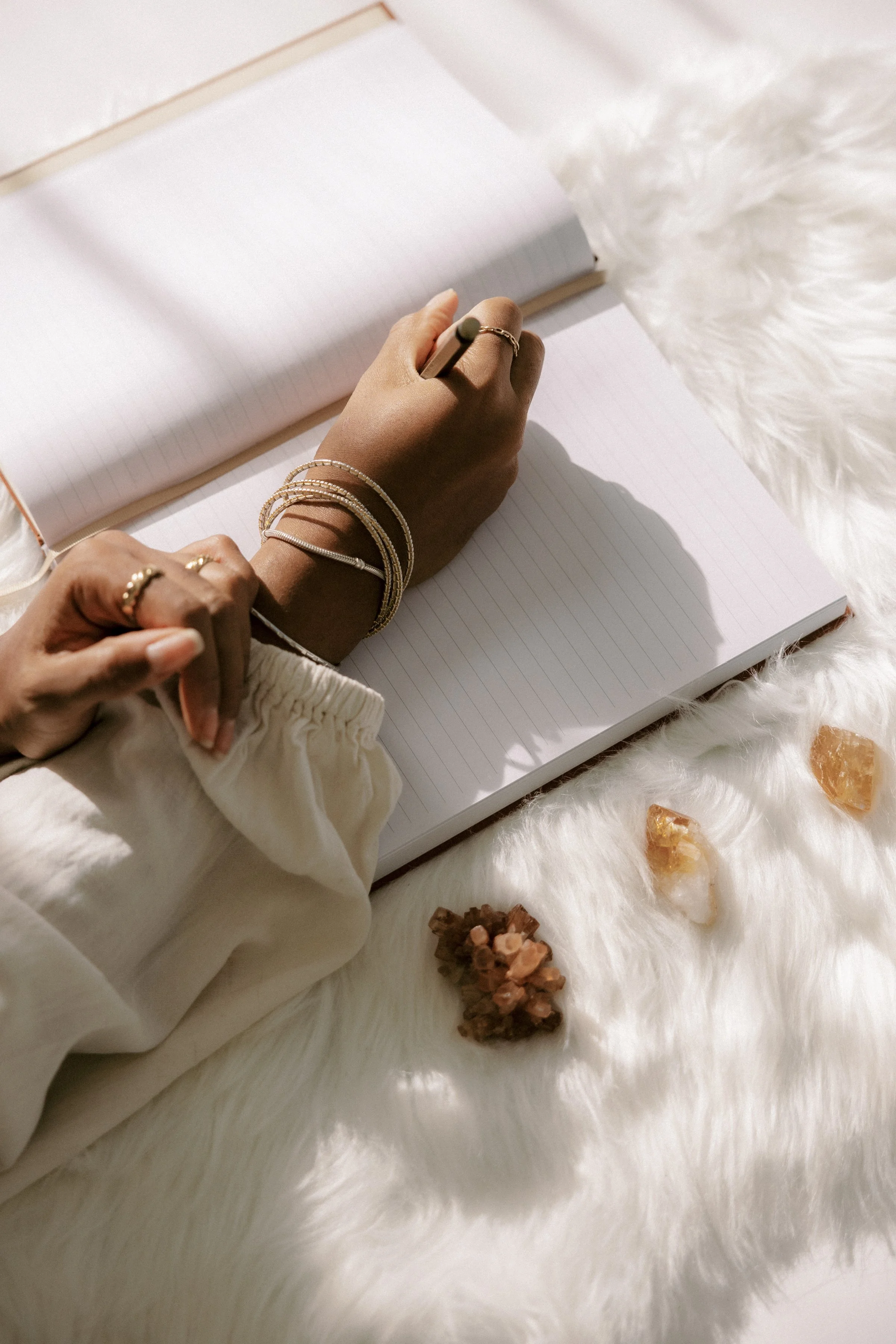 Person’s hand writing in a blank, lined notebook with a gold pen, resting on a soft, white, fluffy surface with small crystals around.