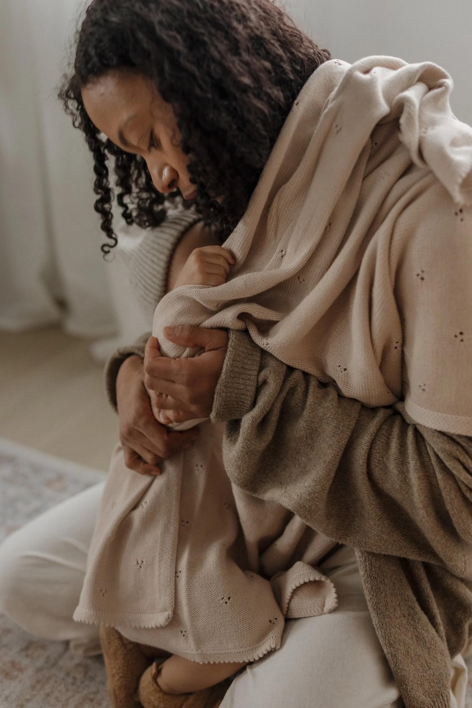 A woman with curly dark hair, wearing a beige hoodie, sitting and holding a small dog or puppy, with her eyes closed and a peaceful expression.