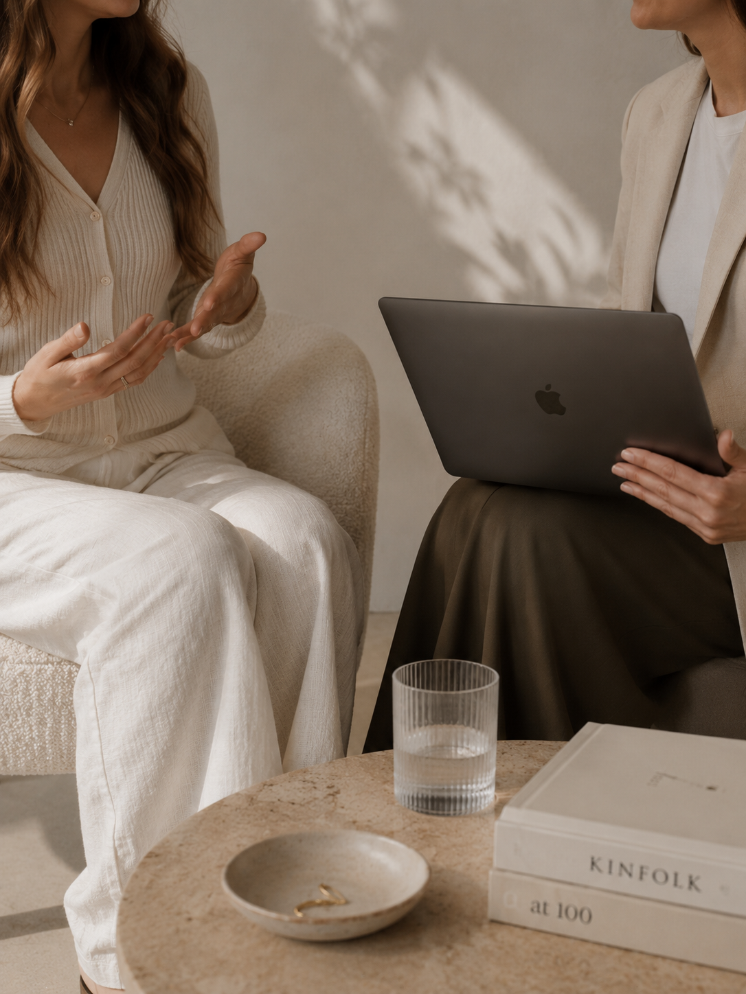 Two women having a conversation in a cozy, neutral-toned room. One woman, wearing a cream cardigan, is gesturing with her hands. The other woman, dressed in a beige blazer and dark skirt, holds a black MacBook laptop. On the table in front of them, there is a glass of water, a ceramic bowl with a gold ring inside, and a book titled 'KINFOLK at 100'.