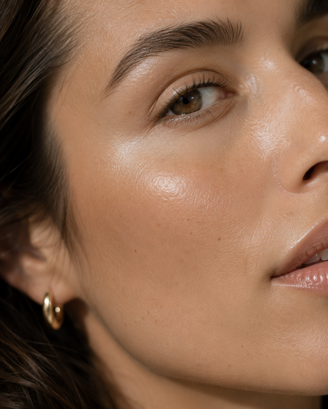 Close-up of a woman's face, highlighting her brown eye, eyebrow, and smooth skin with minimal makeup and a small gold hoop earring.