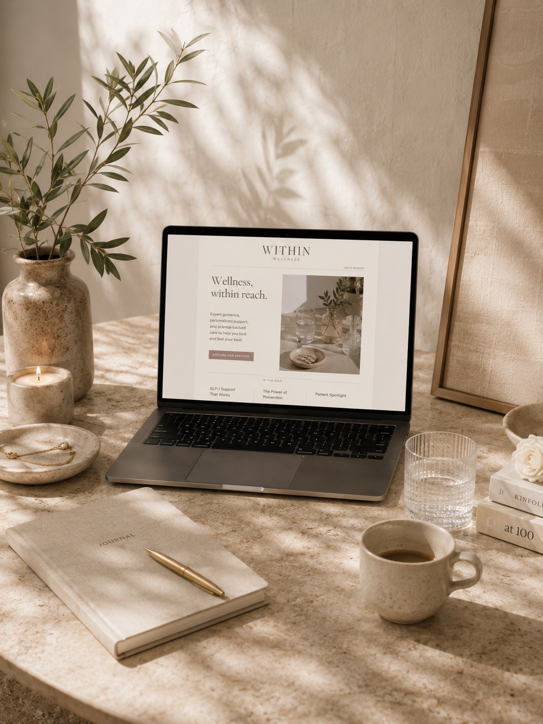 A workspace with a laptop displaying a wellness website, a beige notebook with a gold pen, a coffee mug, a glass of water, a potted plant, a lit candle, a decorative plate, a small dish, and a stack of books on a beige marble desk, illuminated by natural light.