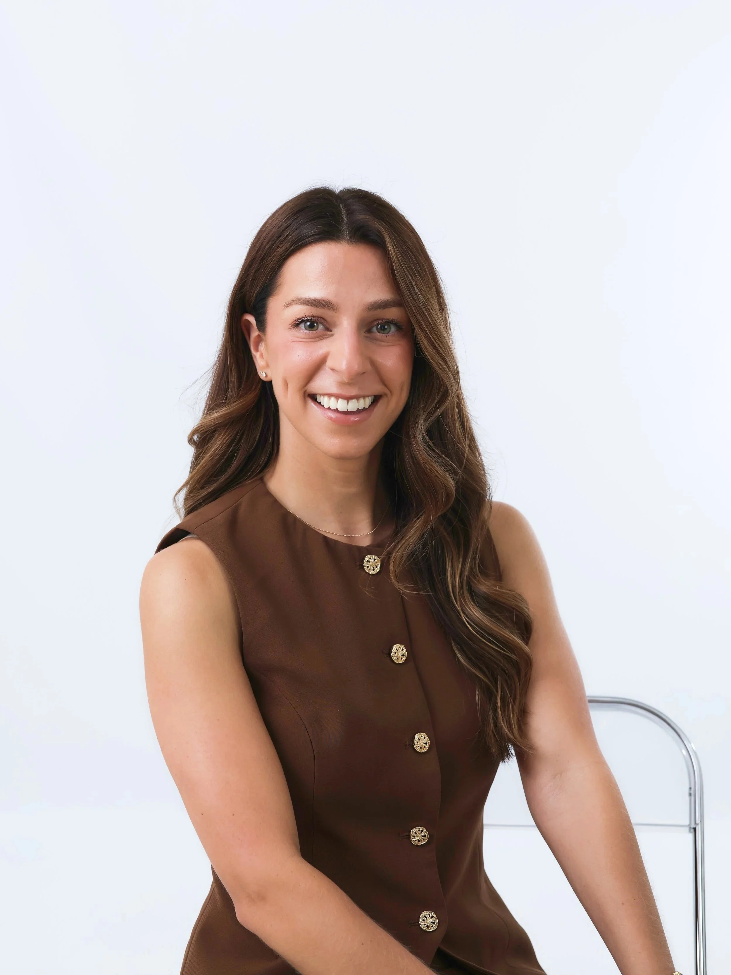 A young woman with long brown hair, smiling, wearing a sleeveless brown dress with decorative gold buttons, sitting on a chair with a metal armrest, against a white background.