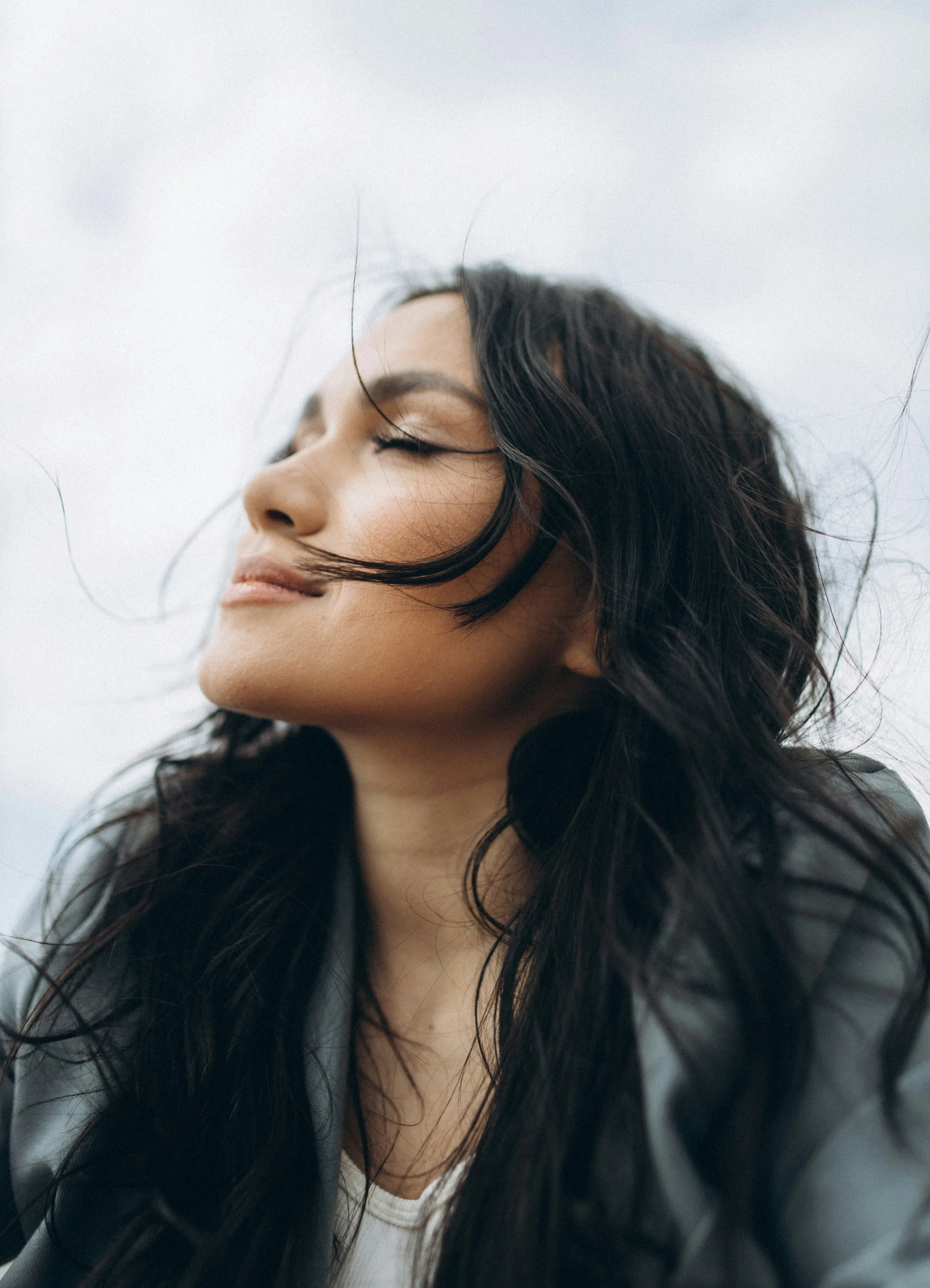 Close-up of a woman with long wavy dark hair and light makeup, with her eyes closed and wind blowing through her hair, against a cloudy sky.