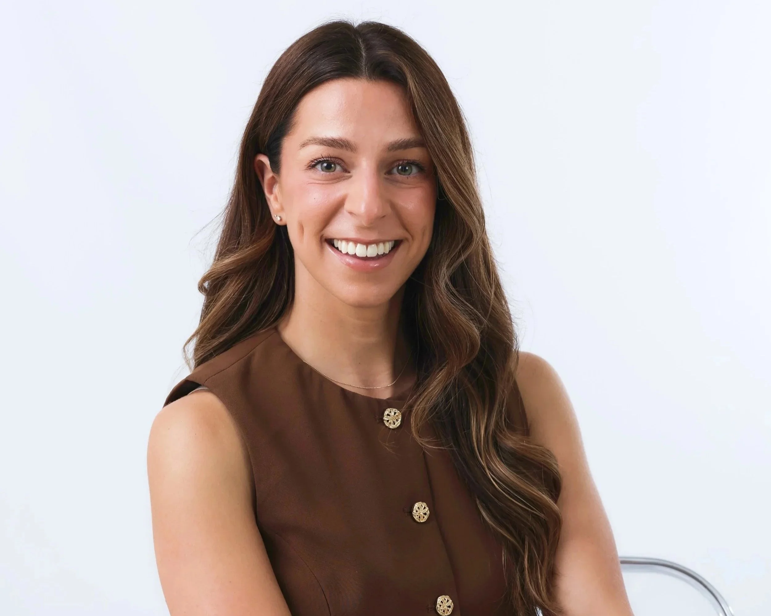 A woman with long brown hair styled in loose curls, smiling, wearing a sleeveless brown top with decorative buttons, standing against a light background.