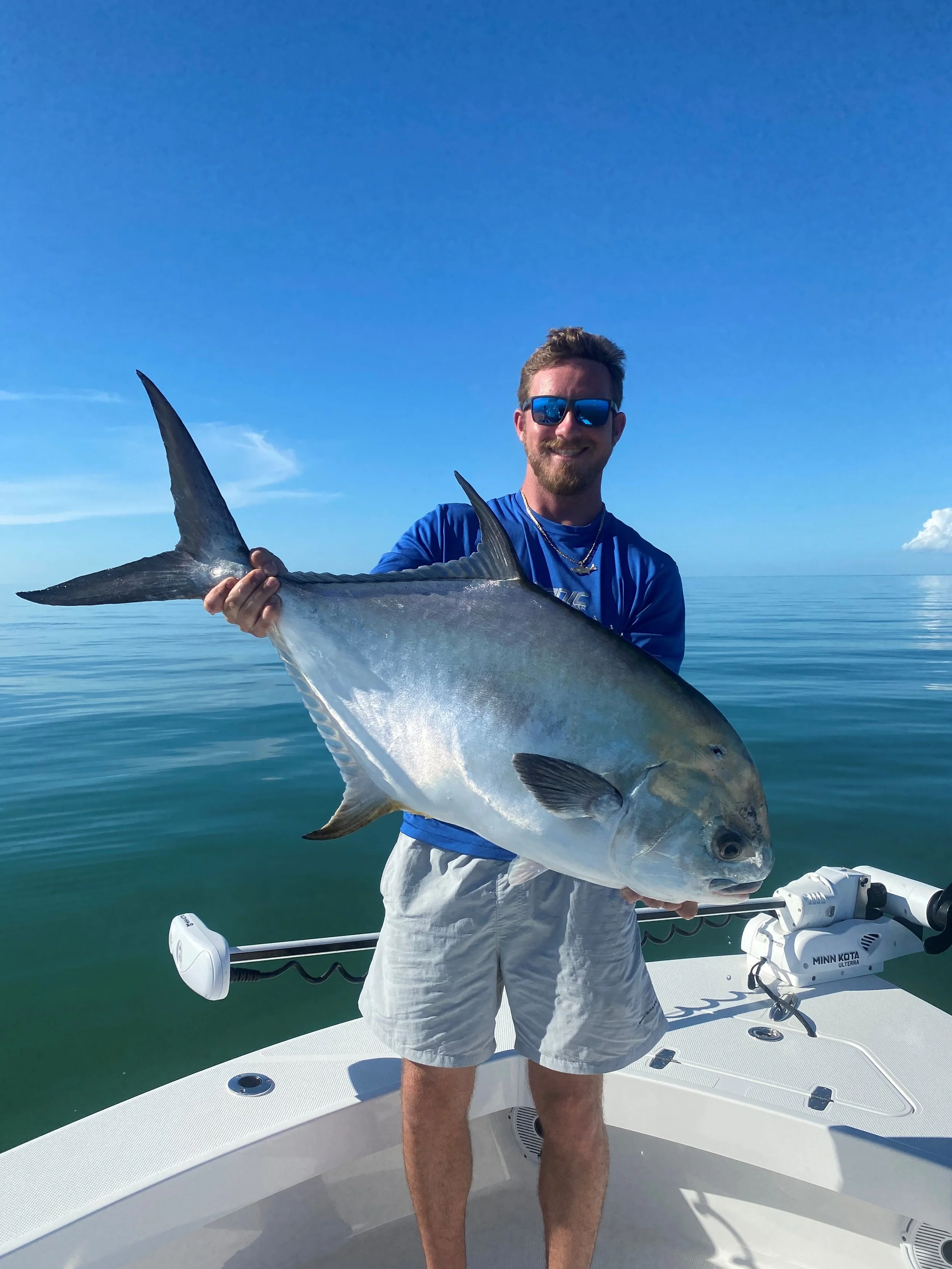 A man wearing sunglasses and a blue shirt stands on a boat, holding a large fish with a silver body and dark tail. The boat is on calm water with a clear blue sky in the background.