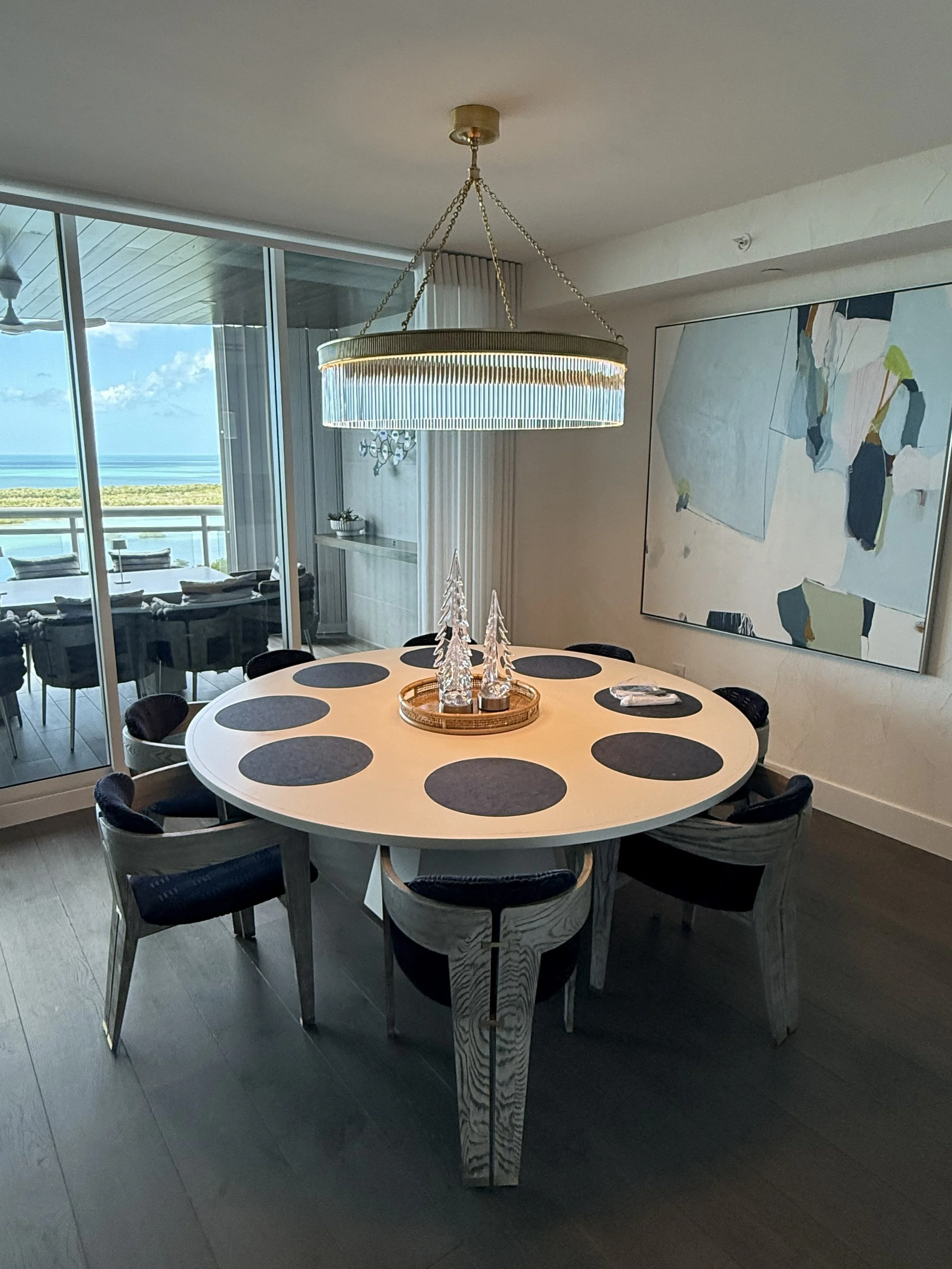 A round dining table with black placemats, surrounded by black chairs, in a modern living room with large window showing an ocean view, artwork on the wall, and a chandelier hanging above.