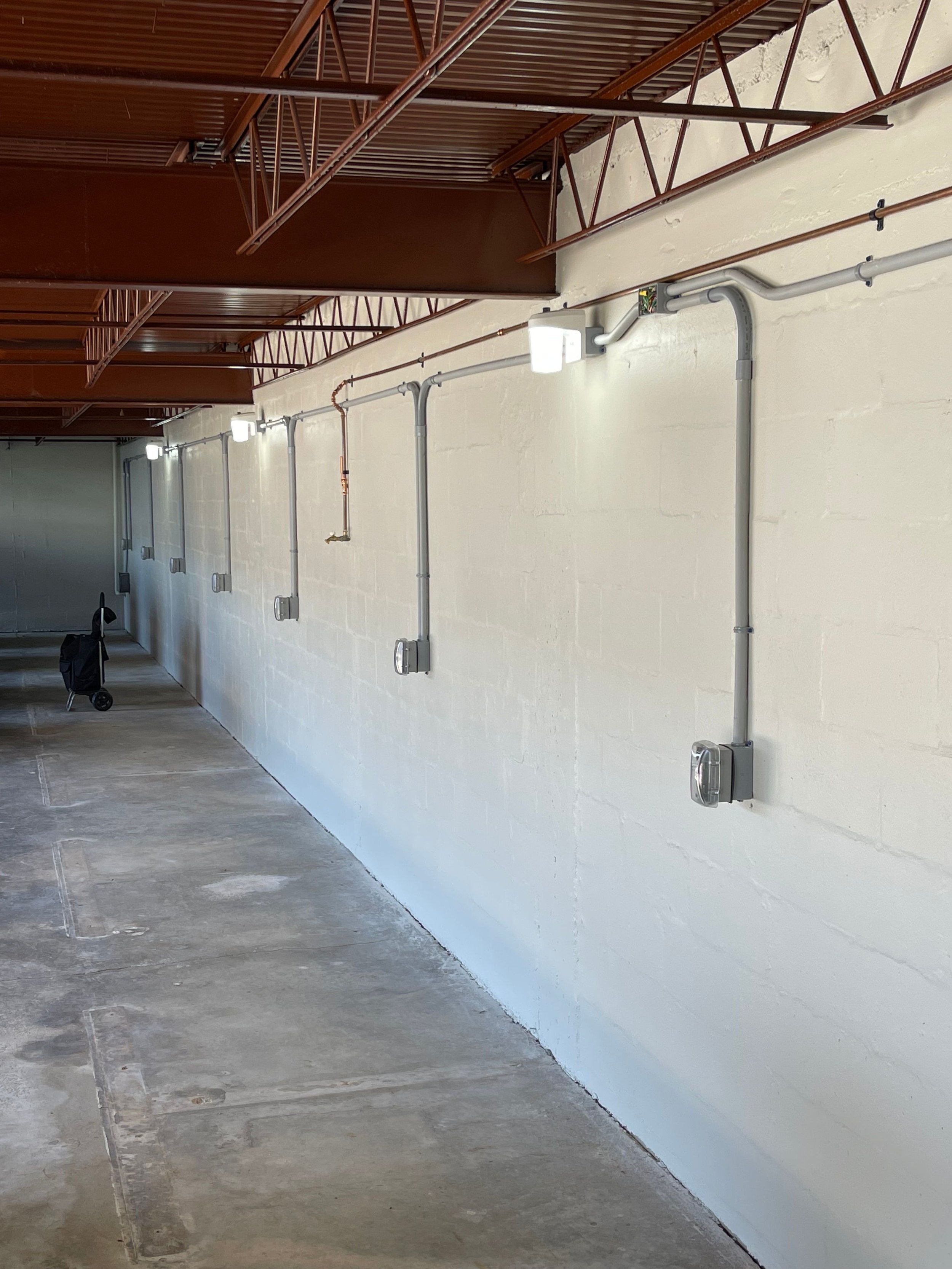 Long white wall with electrical conduit pipes and gray electrical boxes, with brown metal shelving units above. The floor is gray concrete.