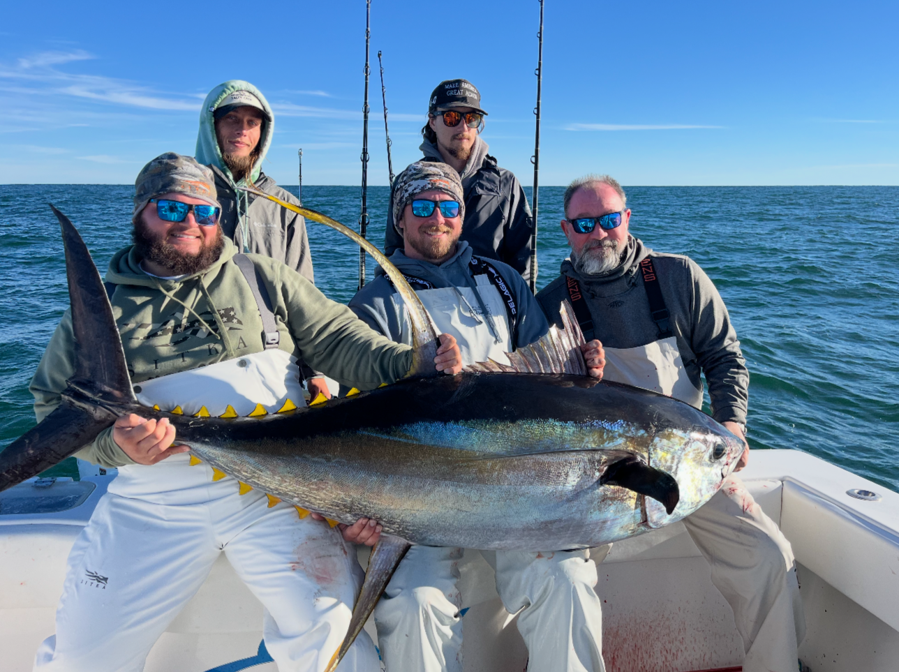 Five men on a boat holding a large fish they caught during a fishing trip, with the ocean in the background.