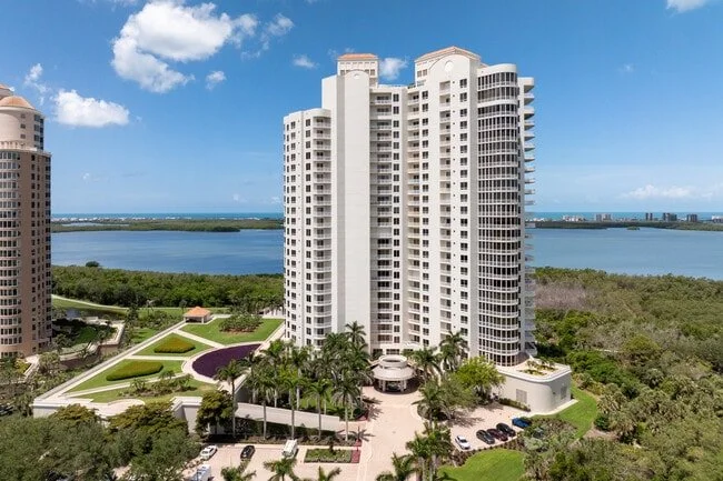 Tall white residential building with balconies, surrounded by green trees, near water with a clear blue sky.