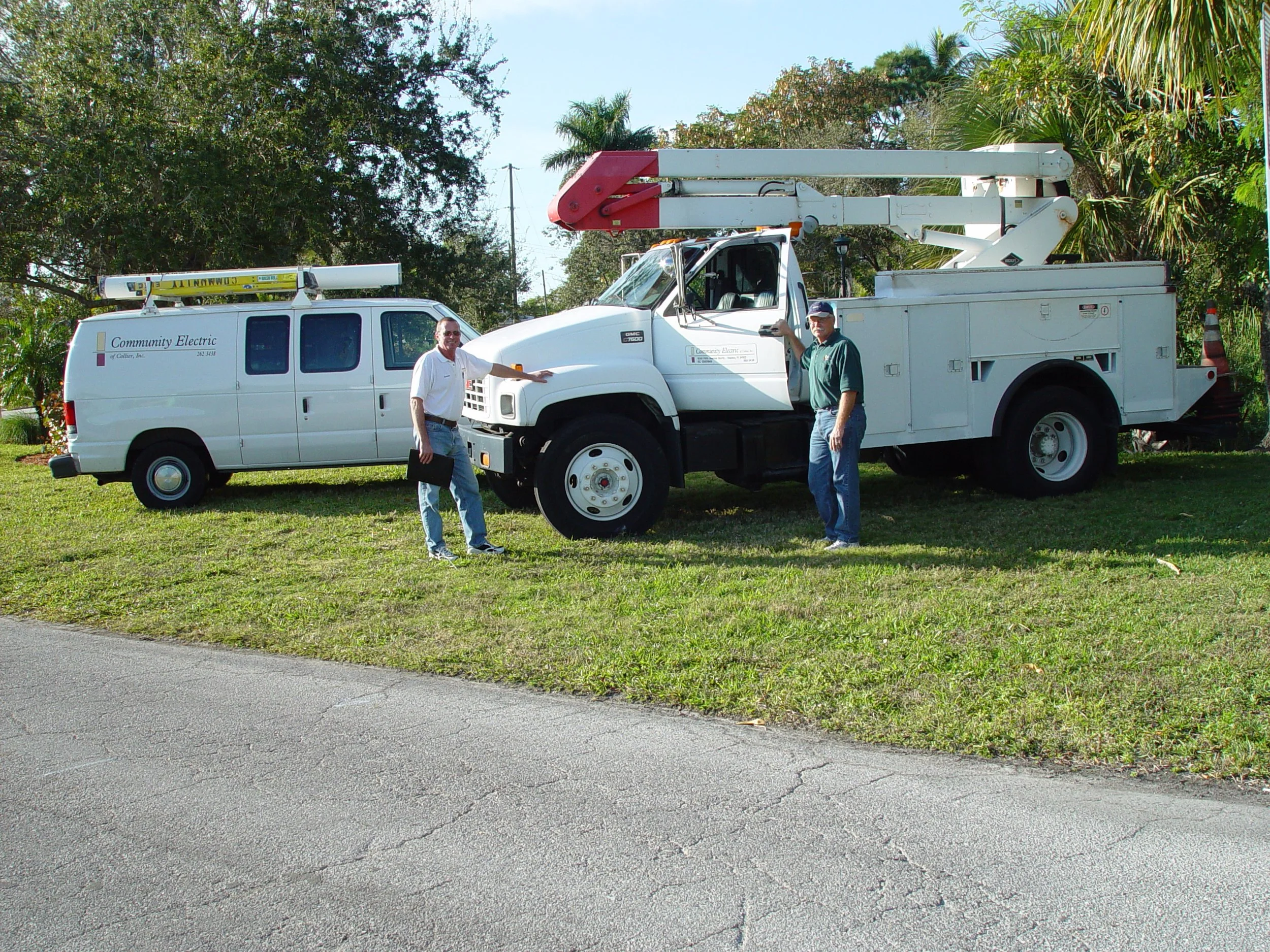 A white utility truck with a bucket lift is parked on grass next to a sidewalk. Two men are standing beside it, smiling and pointing toward the vehicle, with trees and blue sky in the background.