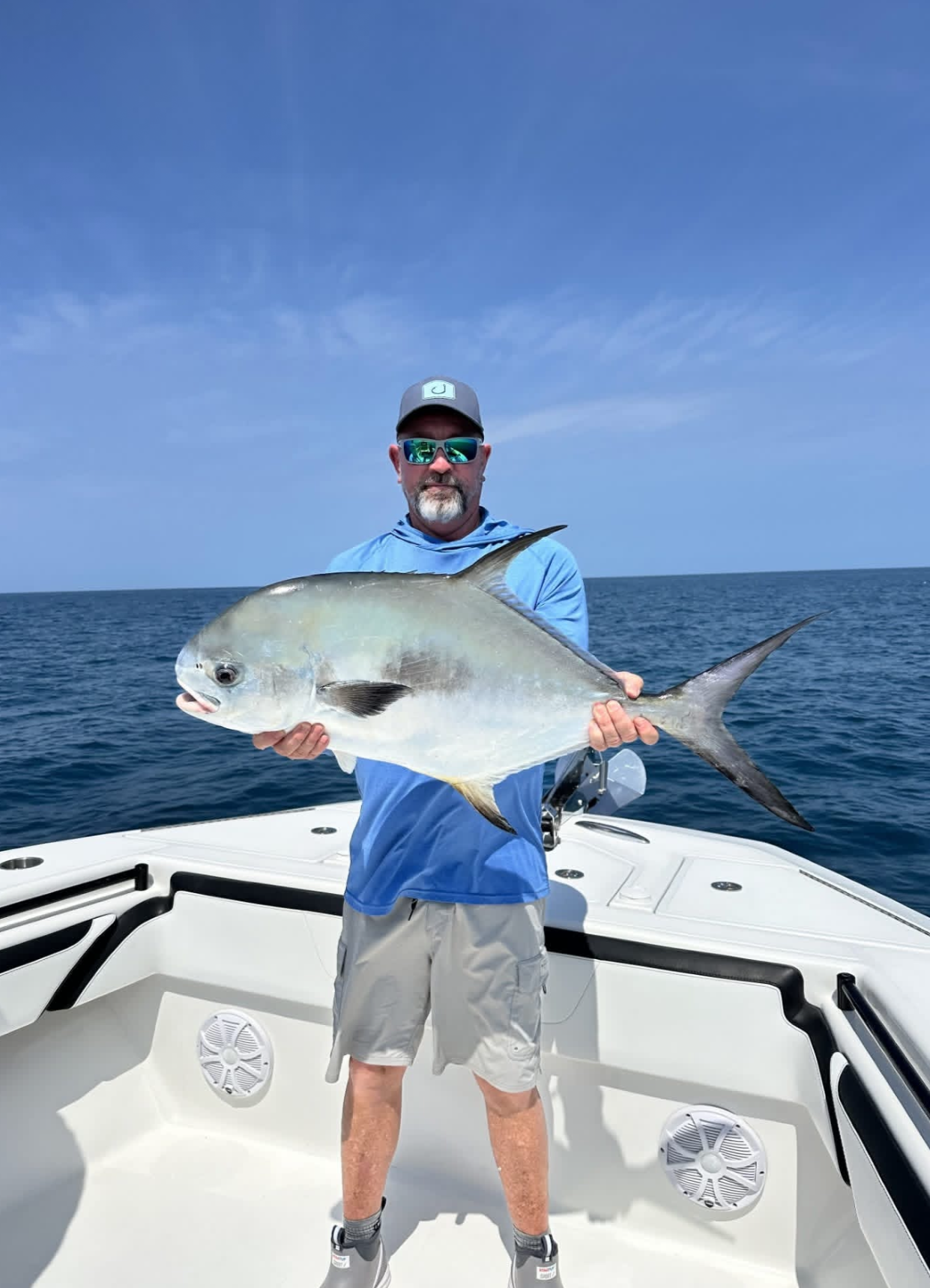 A man in a blue shirt, gray shorts, sunglasses, and a cap is holding a large fish on a boat in the middle of the ocean under a blue sky.