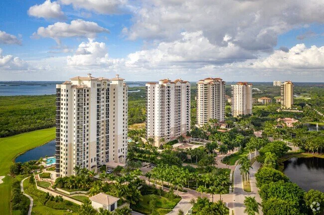 View of a high-rise apartment complex surrounded by lush greenery, with a man-made pond and golf course nearby, under a partly cloudy sky.