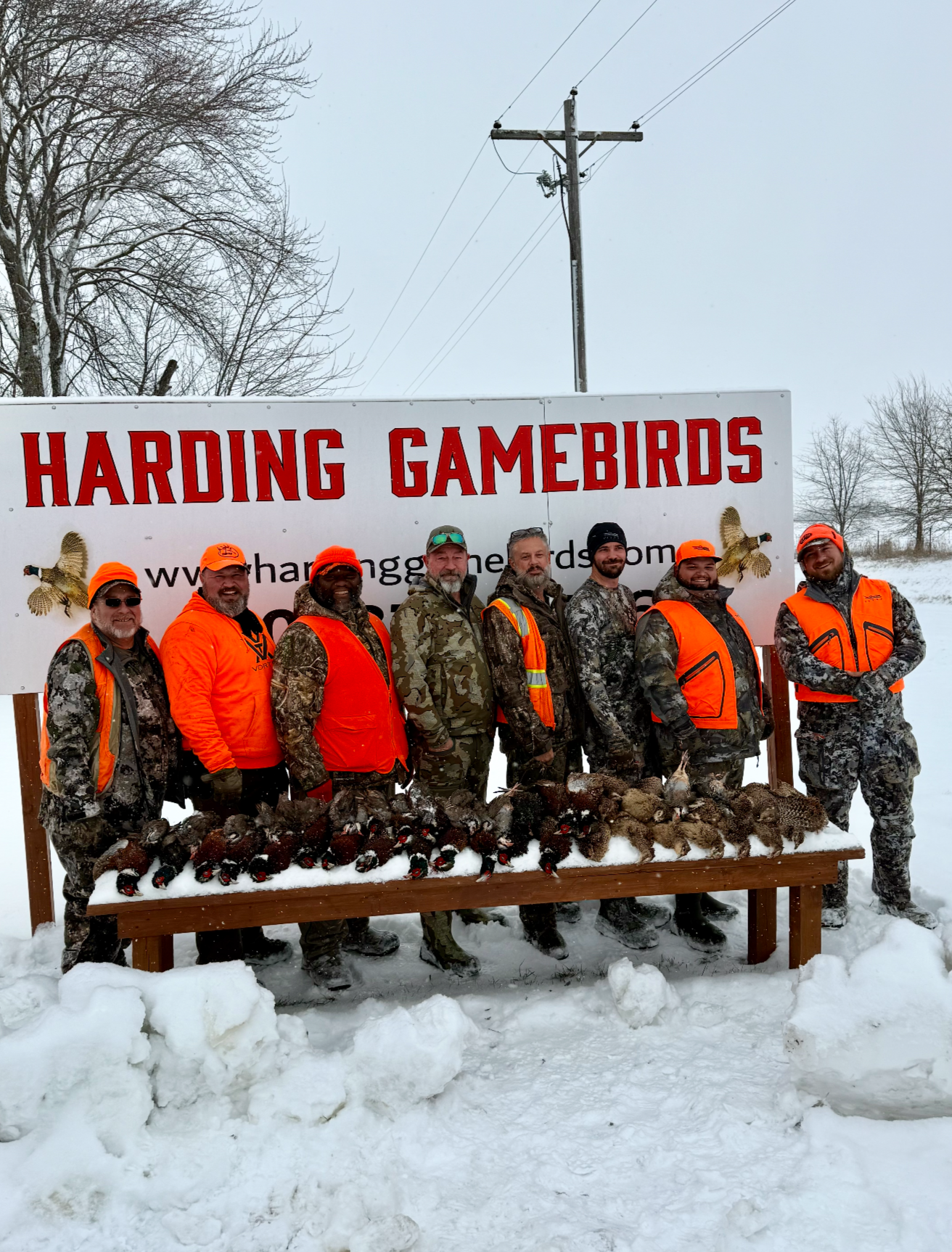 Group of hunters in camouflage and orange safety gear standing behind a table of ducks and turkeys, outdoors in a snowy area, in front of a sign that reads 'HARDING GAMERBIRDS'.