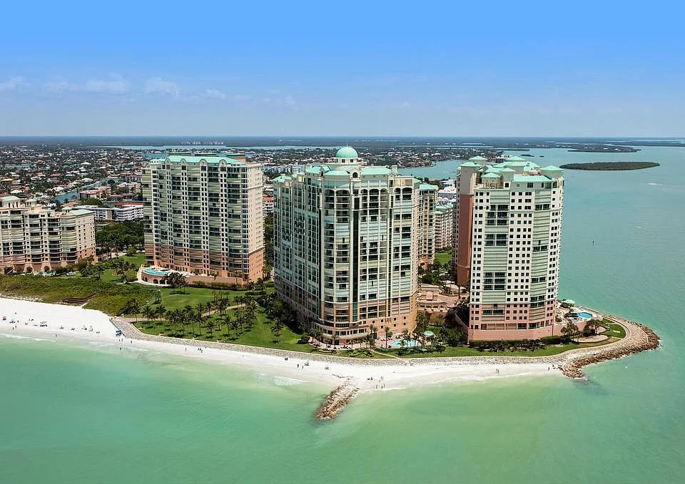 High-angle view of beachfront buildings on an island, with a sandy beach and clear water in the foreground, and a distant cityscape and waterway in the background under a blue sky.