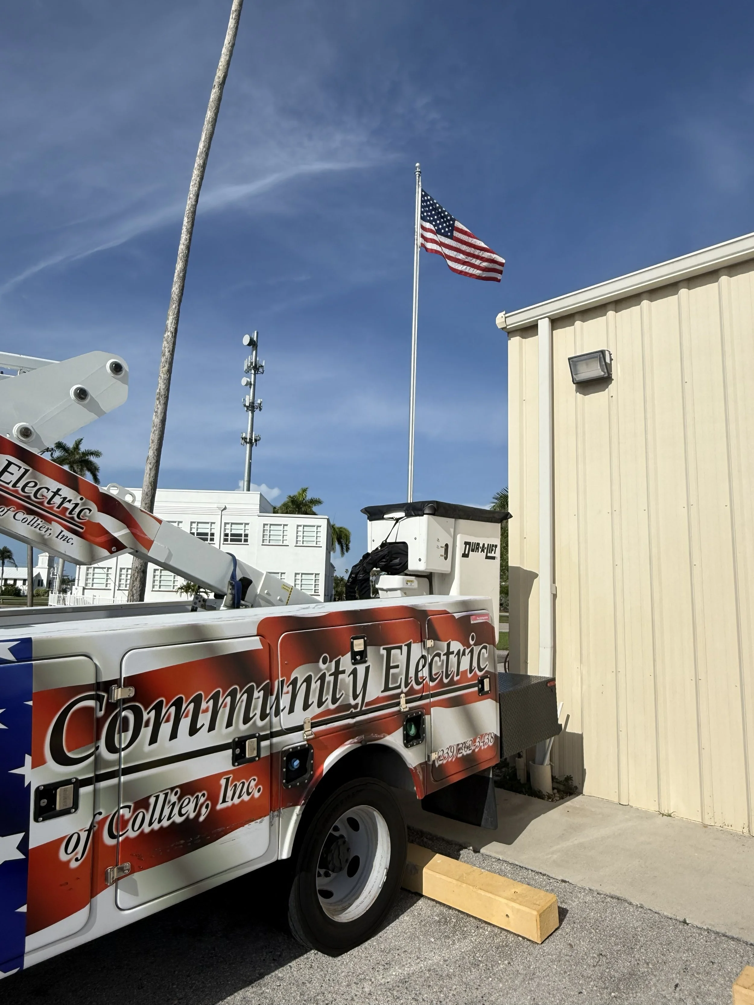 A utility bucket truck with the text 'Community Electric of Collier, Inc.' on the side is parked in front of a beige building. An American flag is flying on a flagpole nearby under a blue sky with some clouds.