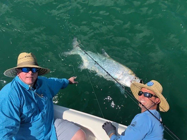 Two men on a boat, holding a large fish that they caught, with green water in the background.