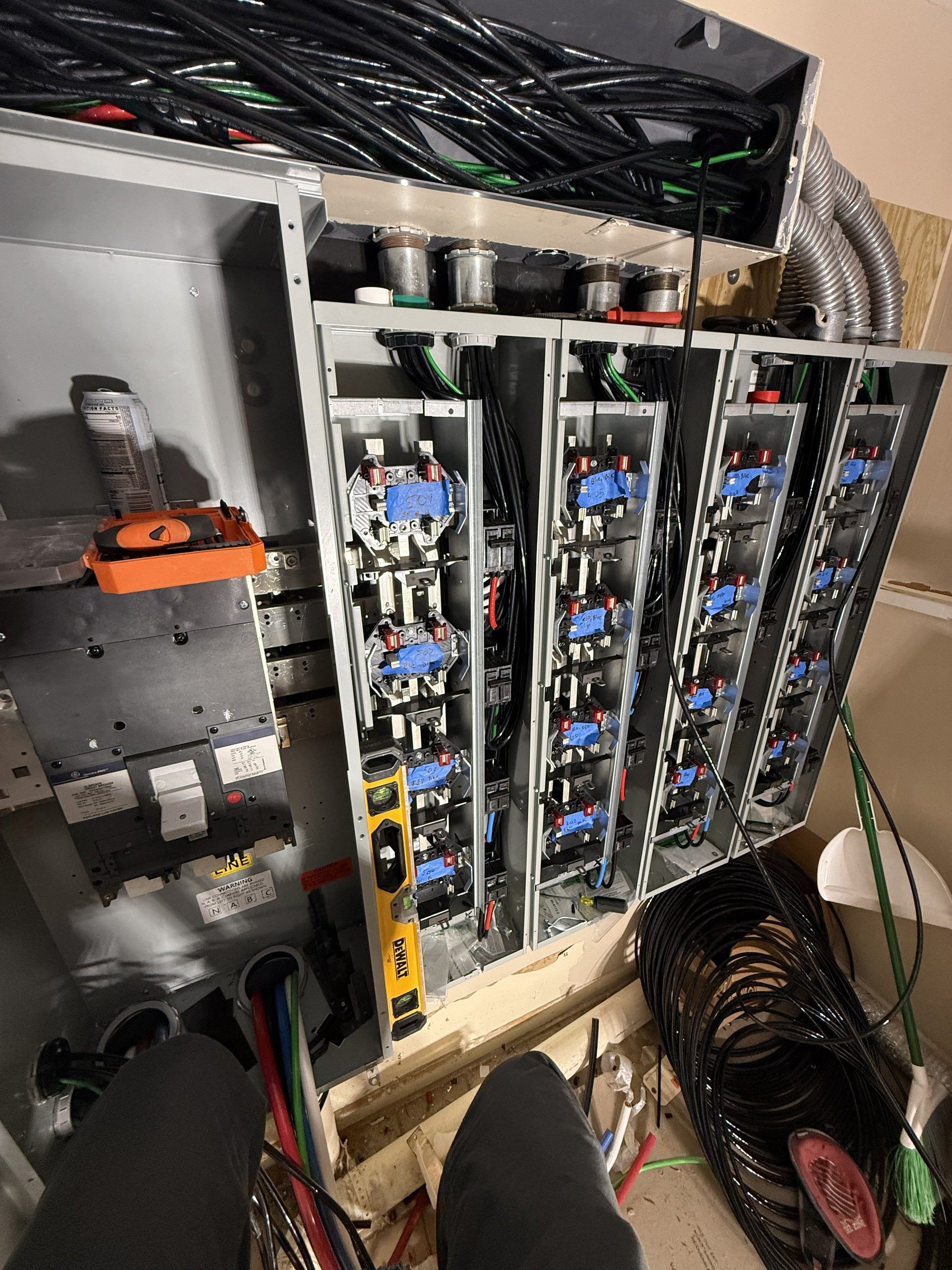 The image shows an electrical panel or control box with multiple circuit breakers or switches, with a person's legs visible at the bottom, and various cables and tools around it, indicating ongoing electrical work.