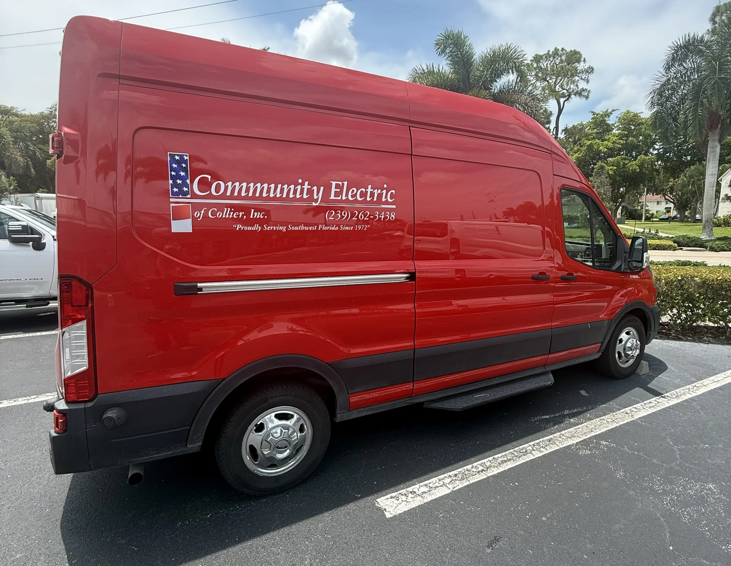A red service van for Community Electric of Collier, Inc. parked in a parking lot with palm trees and residential houses in the background.