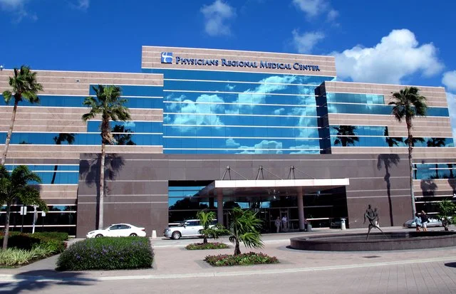 Exterior of the Physicians Regional Medical Center building with palm trees, parking lot, and a large glass window reflecting clouds