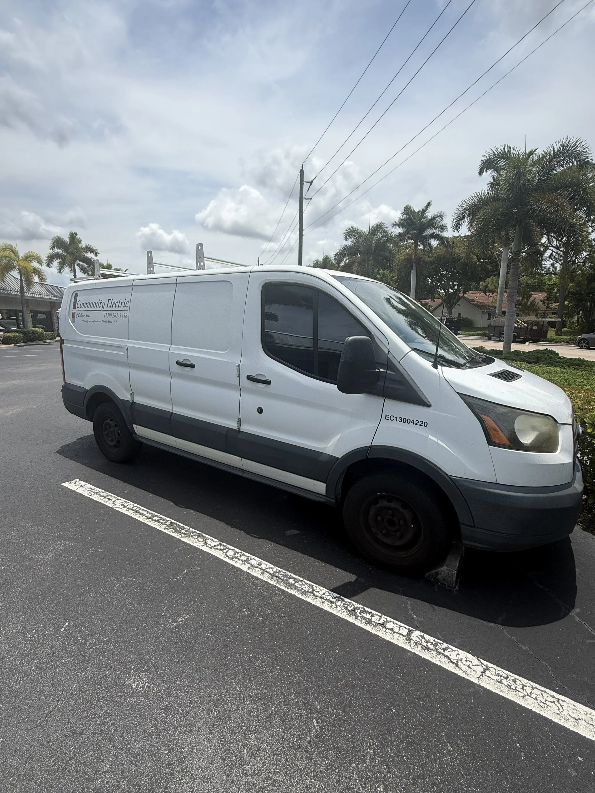 A white service van parked in a parking lot with palm trees and buildings in the background under a partly cloudy sky.