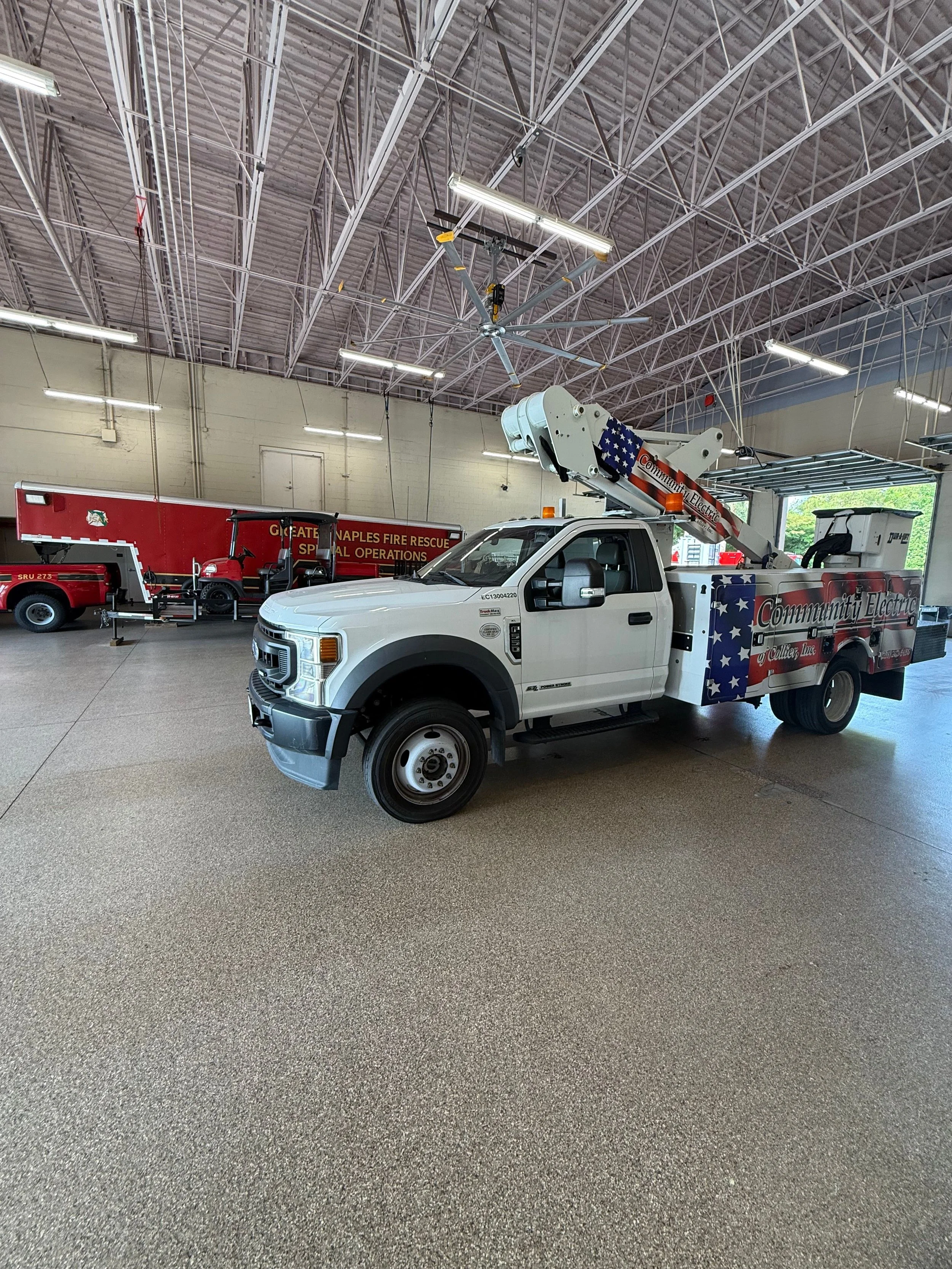 Indoor fire station with emergency vehicles, including a service truck with an aerial lift and a golf cart, under a high ceiling with metal trusses.